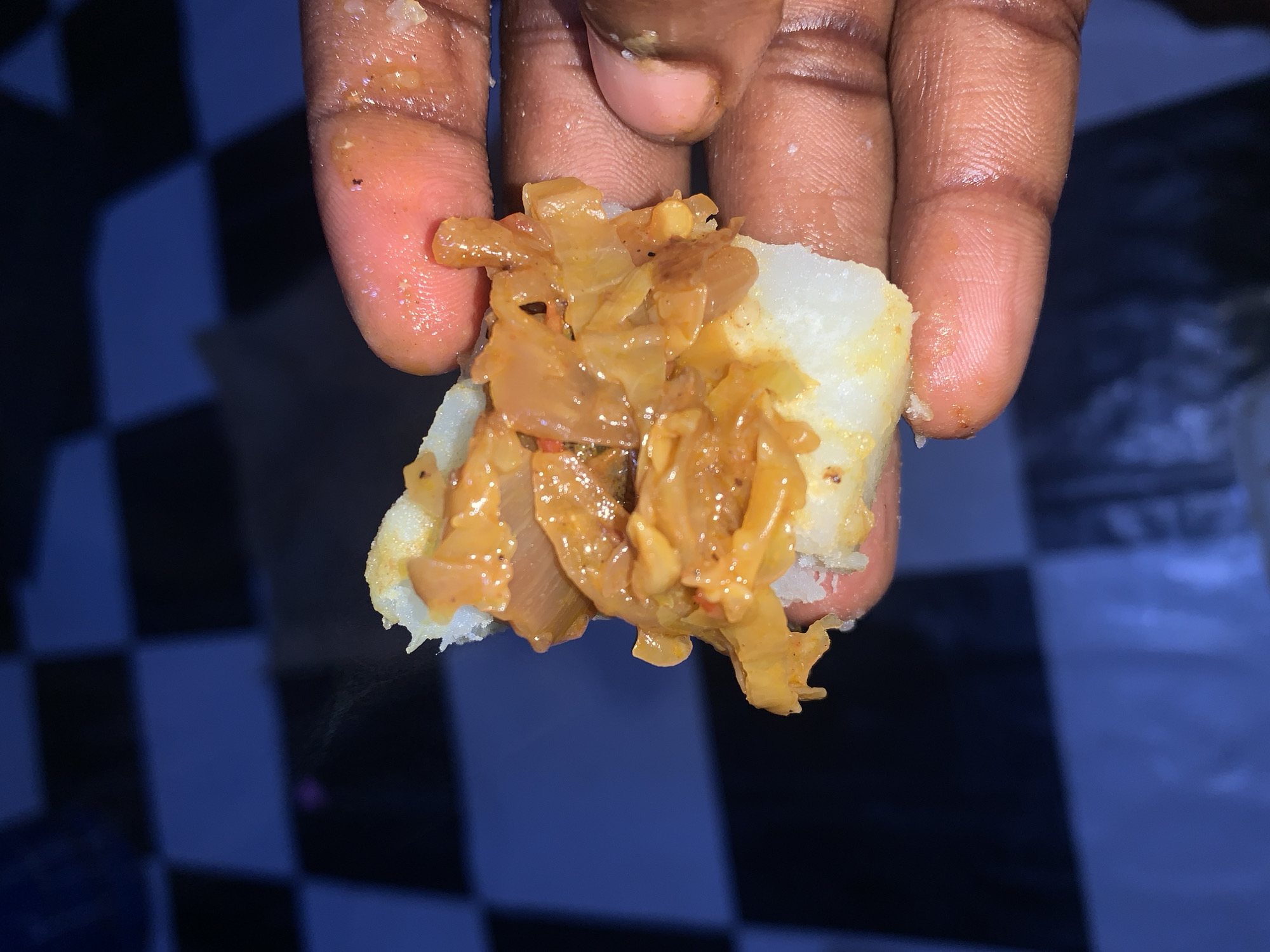 A close-up, high-angle shot shows a person's hand holding a piece of food. The food consists of a white, starchy base topped with shredded, cooked onions in a reddish-brown sauce. The background is out of focus, revealing a dark, checkered pattern that could be a floor or table, and hints of blue. The image is taken in Rafin Par, Nigeria.