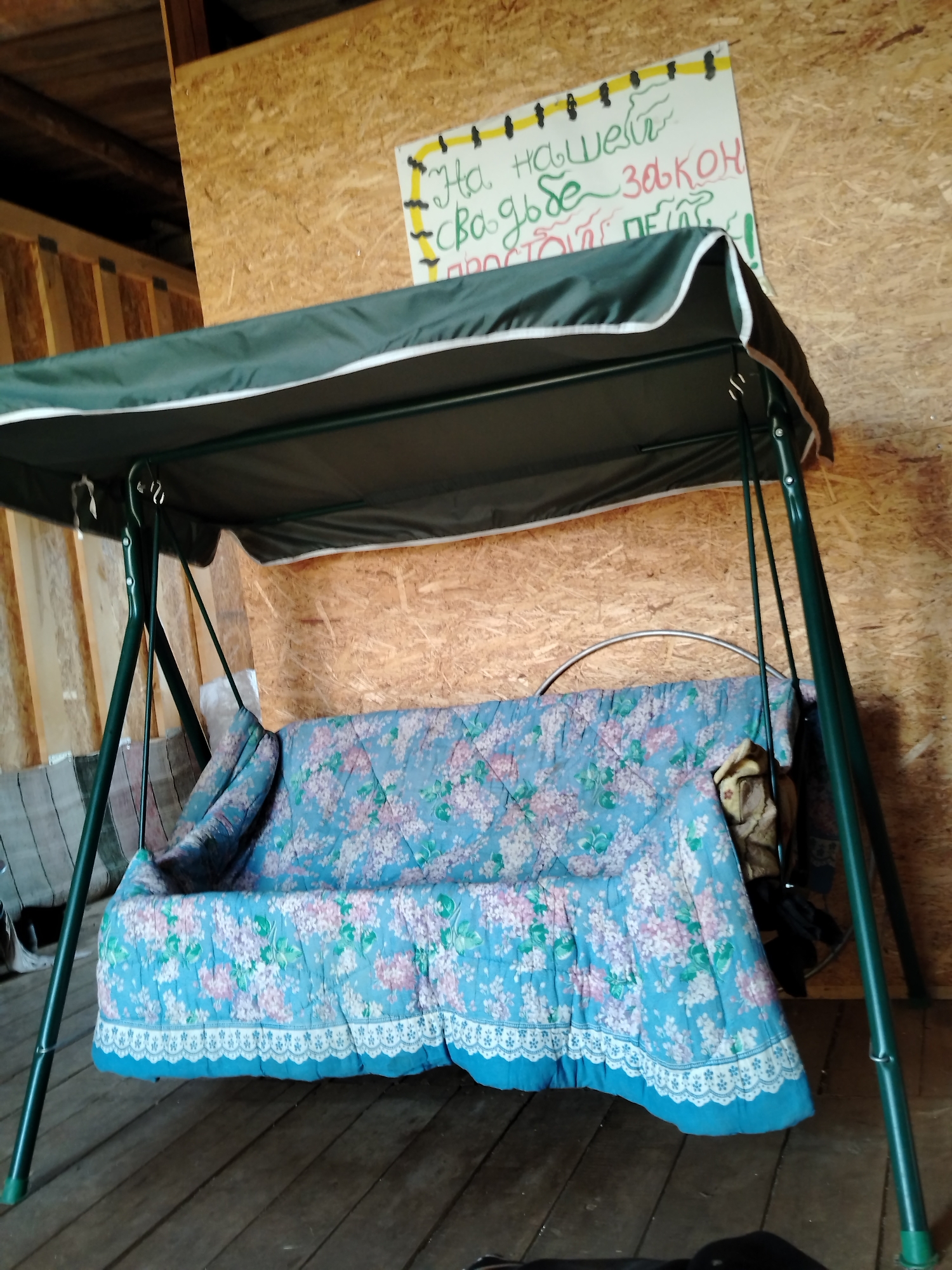The image depicts an indoor scene, possibly a shed or a recreational area, with a swing set as the central object. The swing set is dark green with a dark green canopy and a light blue cushion adorned with a floral pattern of pink and white flowers. The background features wooden walls and a wooden floor. A sign with handwritten Russian text is mounted on the wall behind the swing set. The text is partially visible and appears to be a humorous or informal statement. The overall impression is one of a casual, possibly rustic, setting. The specific location in Rusalevka, Russia is not visually evident from the image itself and would require external information to confirm.