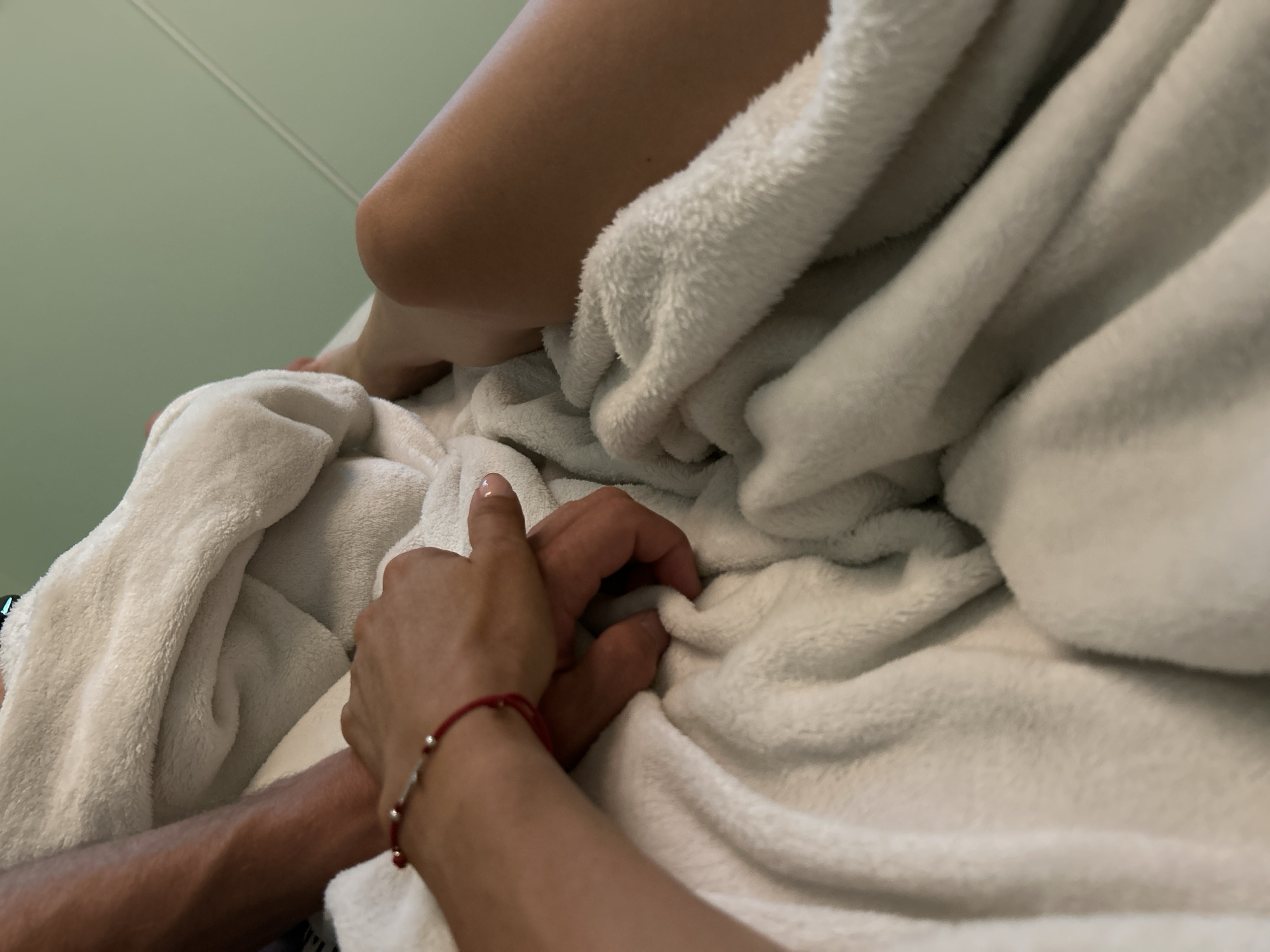 The image is a close-up shot of two people holding hands, partially covered by a white fluffy blanket. The hand on the left, belonging to a person with a red string bracelet, is intertwined with the hand on the right. The background is a pale green wall. There are no discernible landmarks or features that would place this image specifically within the city of Burgas, Bulgaria. The scene appears to be indoors, likely in a domestic setting or a hotel room.