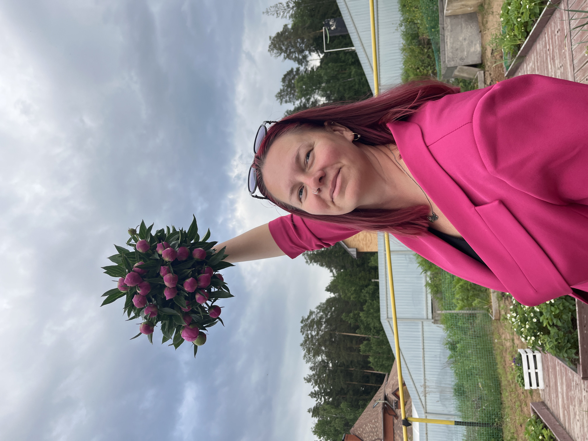 A woman is posing with a bouquet of pink flowers. She is wearing a pink blazer, and sunglasses are atop her head. The location appears to be outdoors, with trees and a cloudy sky in the background. There are buildings in the scene as well. Given the city provided, the location is in Chaikovsky, Russia.