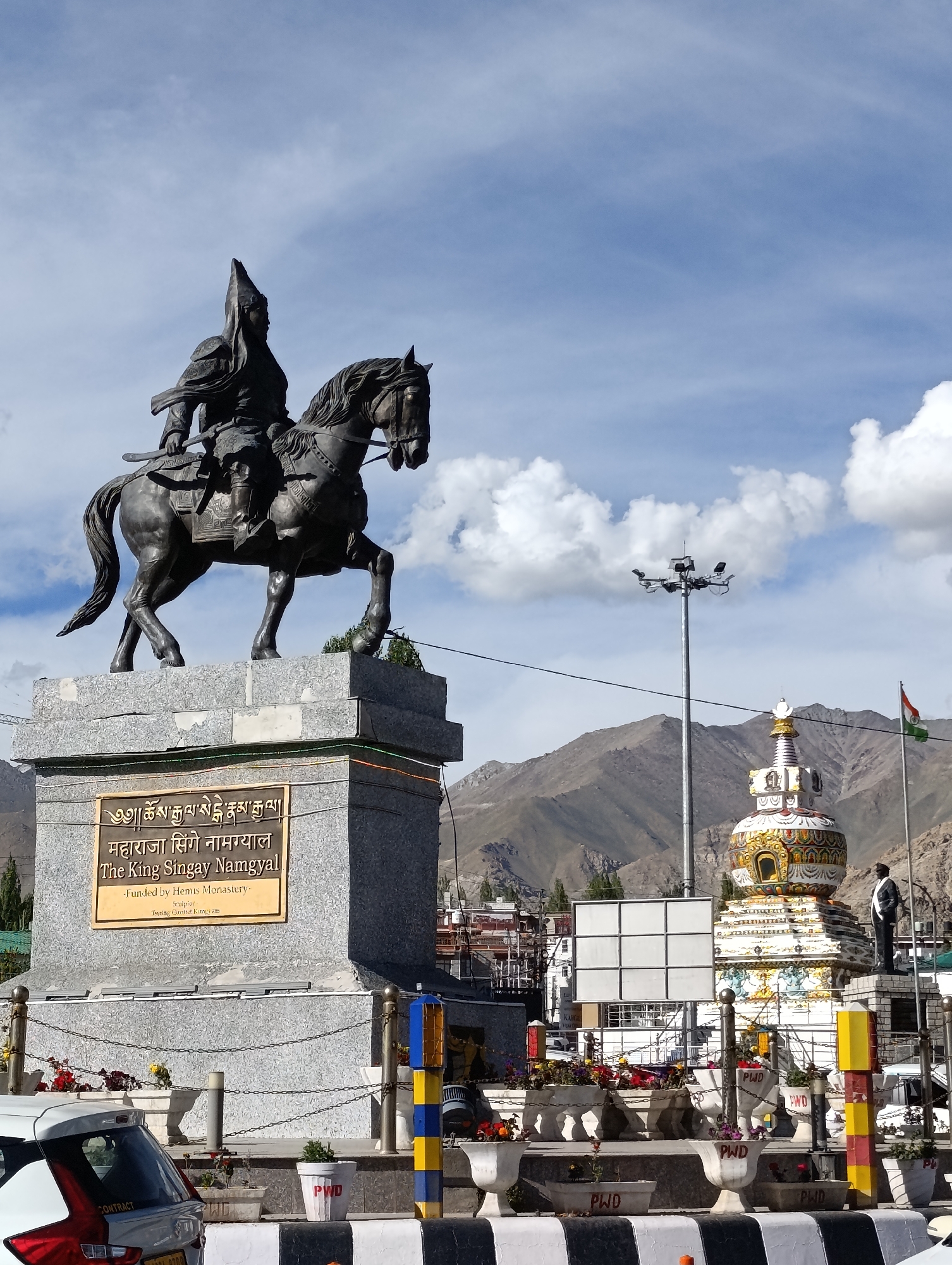 A bronze equestrian statue of King Singay Namgyal stands on a stone pedestal in Leh, India. The king wears traditional attire and a pointed helmet. A golden plaque on the pedestal displays text in Tibetan script and English, identifying him and stating that the statue was funded by Hemis Monastery. Behind the statue, a Tibetan-style stupa with ornate decorations is visible, along with mountains in the background. An Indian flag flies from a flagpole to the right of the stupa. A tall street light with multiple lamps stands to the left of the stupa. In the foreground, a white car is parked, and planters with flowers line a walkway. The sky is blue with scattered white clouds.