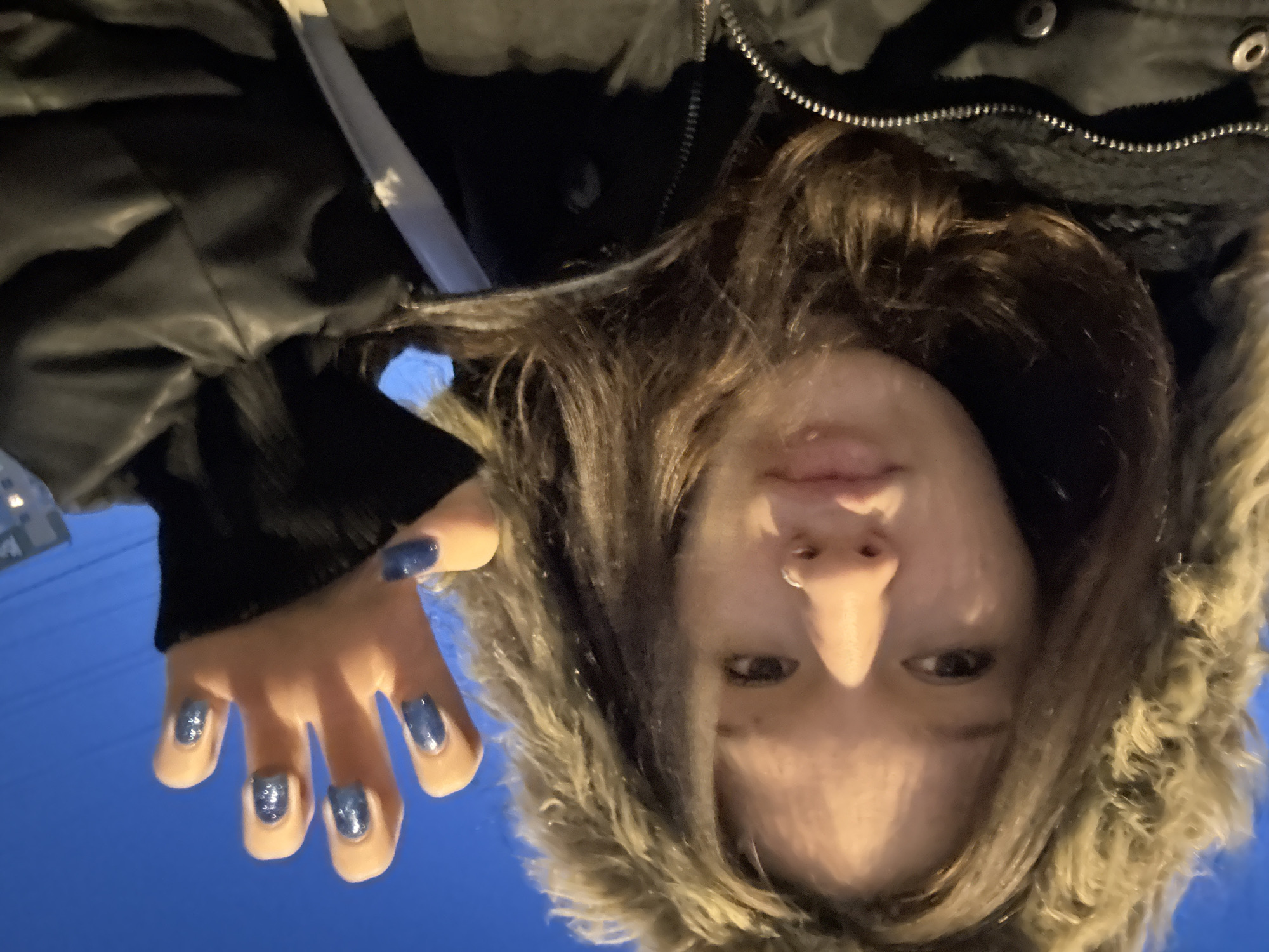 The image is a rotated selfie of a person, likely female, captured outdoors against a deep blue sky, suggesting dusk or night. The person is looking directly at the camera with a neutral expression. She has long, wavy brown hair and is wearing a dark winter coat with a fluffy, light brown fur-lined hood. A subtle septum piercing is visible on her nose. Her left hand is raised towards the camera, showcasing fingernails painted with a sparkling blue glitter polish. The texture of the coat and fur, along with the glitter on the nails, are prominent details. The image is oriented 90 degrees counter-clockwise from a standard upright view. The winter attire implies a cold weather setting. There is no visible text.