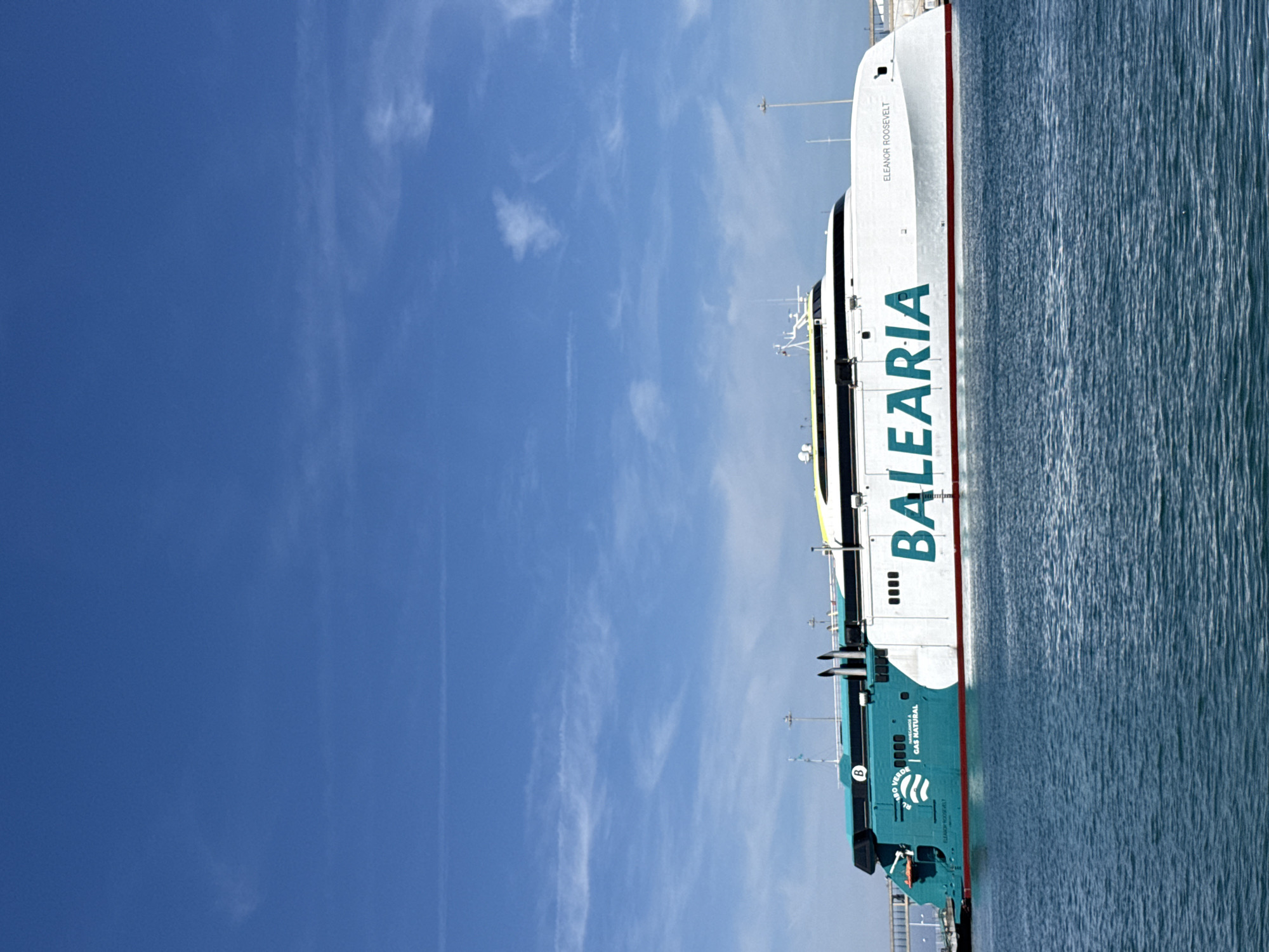 The image depicts a large Balearia ferry docked in Dénia, Spain. The vessel's side is prominently displayed, featuring the company logo and the name "Eleanor Roosevelt" on the upper section. The ferry is situated in a port environment, with calm blue water visible on one side and a clear blue sky with wispy clouds above. The image appears to be taken during the daytime.