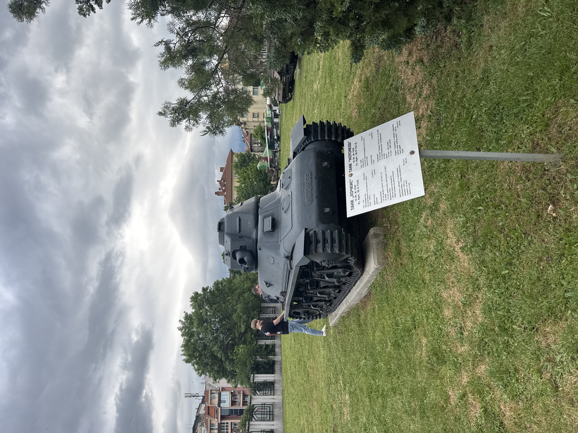 The image shows a Hotchkiss tank, model H-39 (1939), also known as Pz.Kpfz. 38 H 735(f), on display in what appears to be a park or museum. A sign providing technical specifications of the tank is affixed to a pole nearby. A man is leaning on the tracks of the tank, likely looking at the information plate attached to the tank. Buildings are visible in the background, suggesting an urban setting. The sky is cloudy and overcast. The tank sits on a concrete platform within a grassy area. There is a similar tank parked behind. Based on the sign and the Cyrillic lettering, this is located somewhere in Eastern Europe. Sofia, Bulgaria is printed on the sign, indicating the location.
