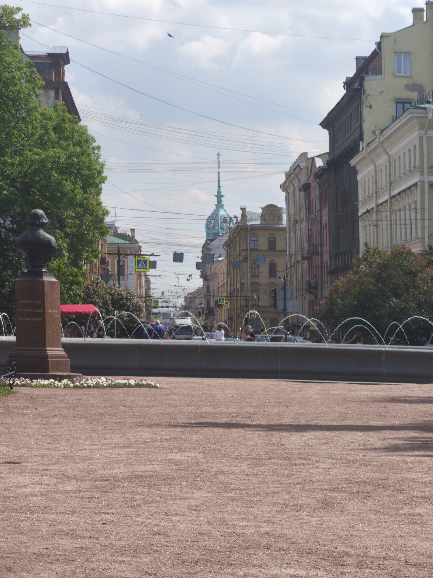 This is a medium shot of a fountain in Saint Petersburg, Russia. The fountain is in the foreground with a statue on the left. Behind the fountain, a street is visible with buildings on either side. People are walking and sitting near the fountain and on the street. The sky is blue with some clouds.
The statue is a bust of a man on a pedestal. The pedestal has inscriptions on it. The fountain is made of stone and has water spraying out of it in arcs. The street is paved with asphalt and has traffic lights and street signs. The buildings are tall and ornate, typical of Saint Petersburg architecture. The trees are green and lush. The overall scene is peaceful and picturesque.
