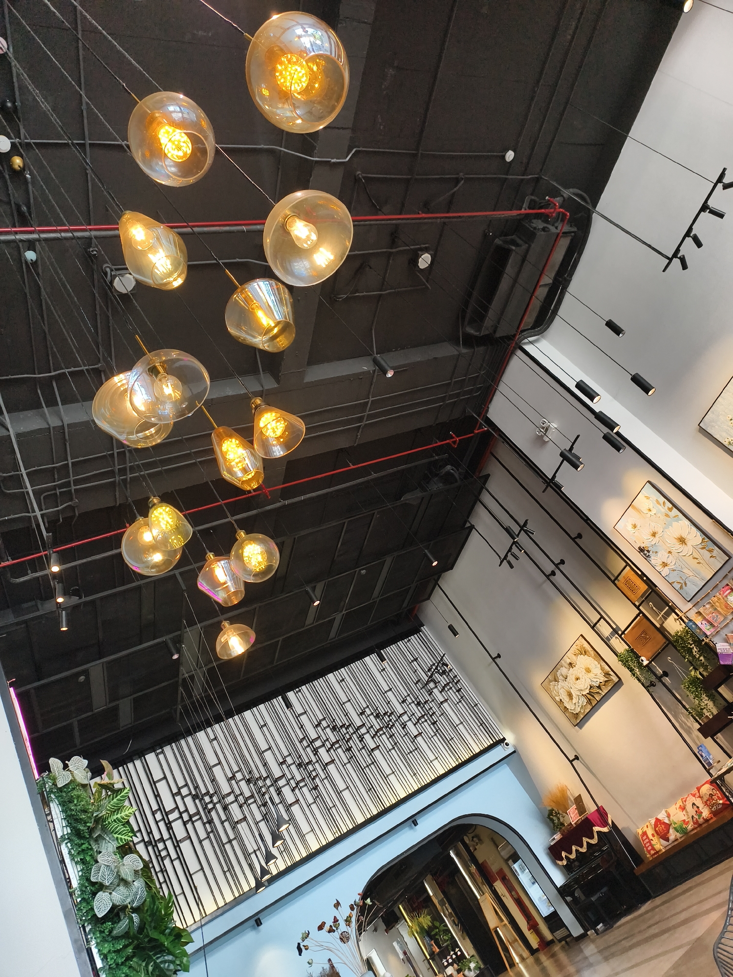 The image captures an interior space with a modern and slightly industrial aesthetic. The ceiling is painted a dark color and features exposed pipes and wiring, with a series of decorative light fixtures hanging down. These lights are of varying shapes and sizes, emitting a warm, golden glow.
To the right, the walls are painted white and adorned with artwork, including framed floral paintings. A series of shelves displays various items, suggesting a retail or display area.
Below the artwork, a large, decorative metal screen is visible, adding to the modern design. The scene transitions into a curved doorway leading to another part of the establishment, possibly a salon or boutique, based on the interior decor and styling. The scene is well-lit, creating a contrast between the dark ceiling and the brighter, more decorated areas below.