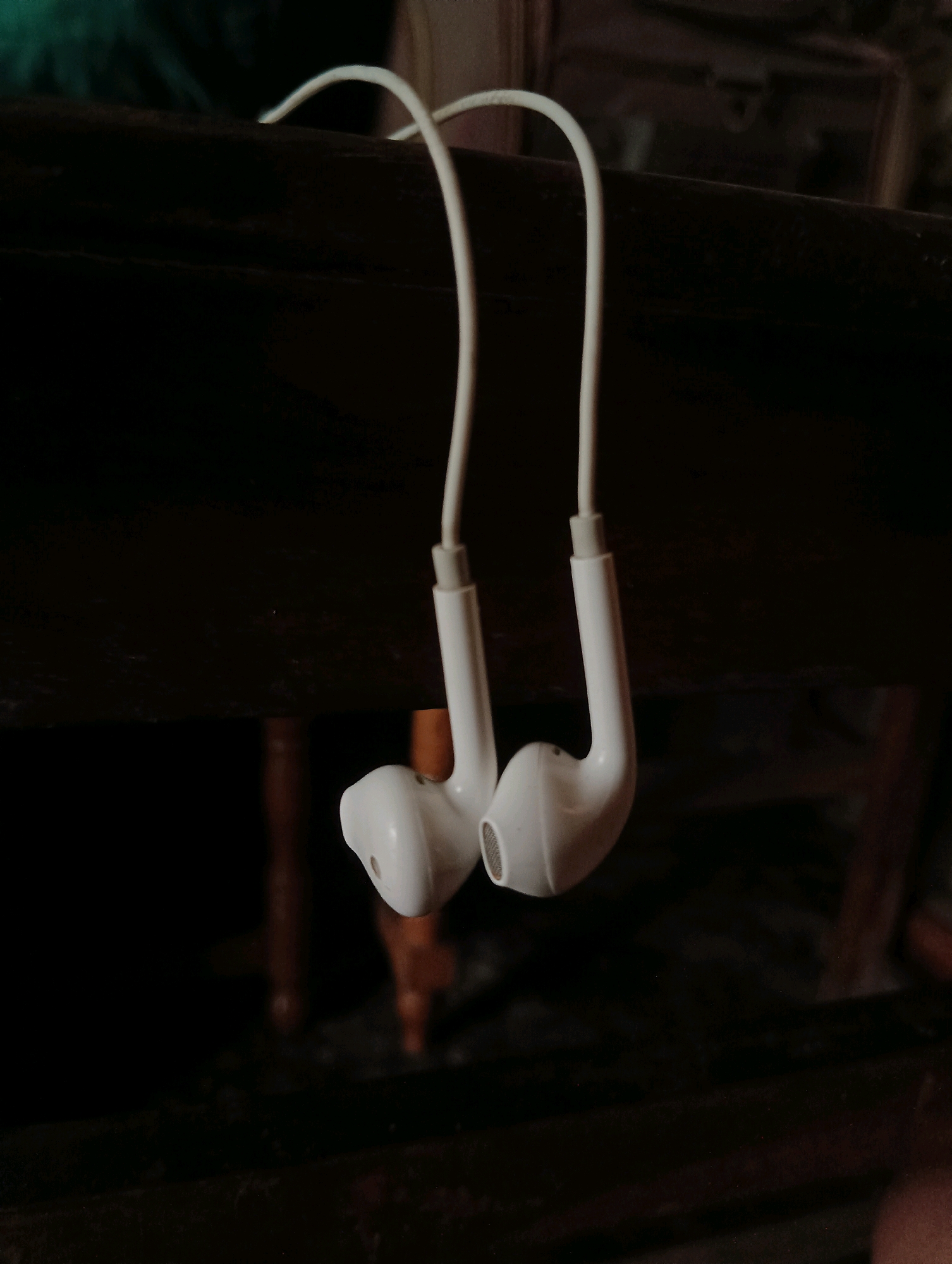 In Dakhin Parsa, India, a pair of white earbuds is hanging by their cables from a dark-colored wooden surface. The earbuds are suspended downwards, with one earbud on the left and the other on the right. The lighting in the scene is dim, casting shadows and highlighting the white color of the earbuds. The background is dark and out of focus, suggesting an indoor setting.