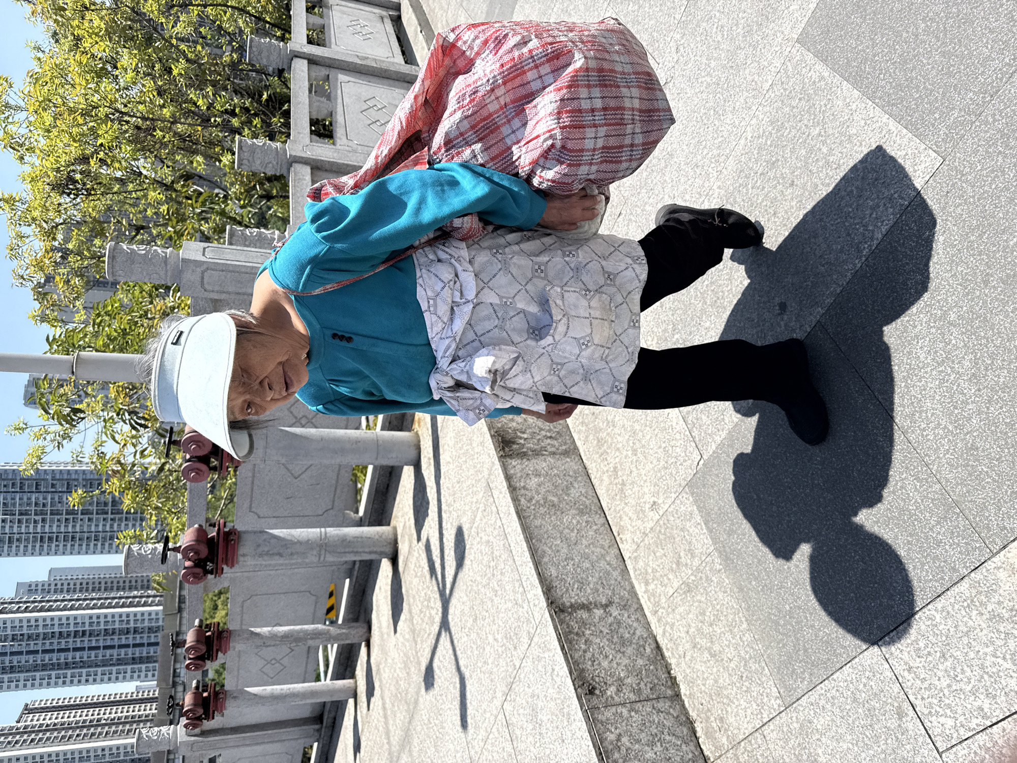 The image shows an elderly woman on a pedestrian walkway in Shapingba District, China. She is wearing a light-colored visor, a blue long-sleeved shirt, black leggings, and black shoes. She has a large red and white checkered bag slung over her shoulder, and another white patterned cloth draped over her shoulders as well. She is walking along a grey stone walkway with shadows. A low stone barrier with metal fixtures is visible to her left, with buildings in the background, indicating an urban setting.