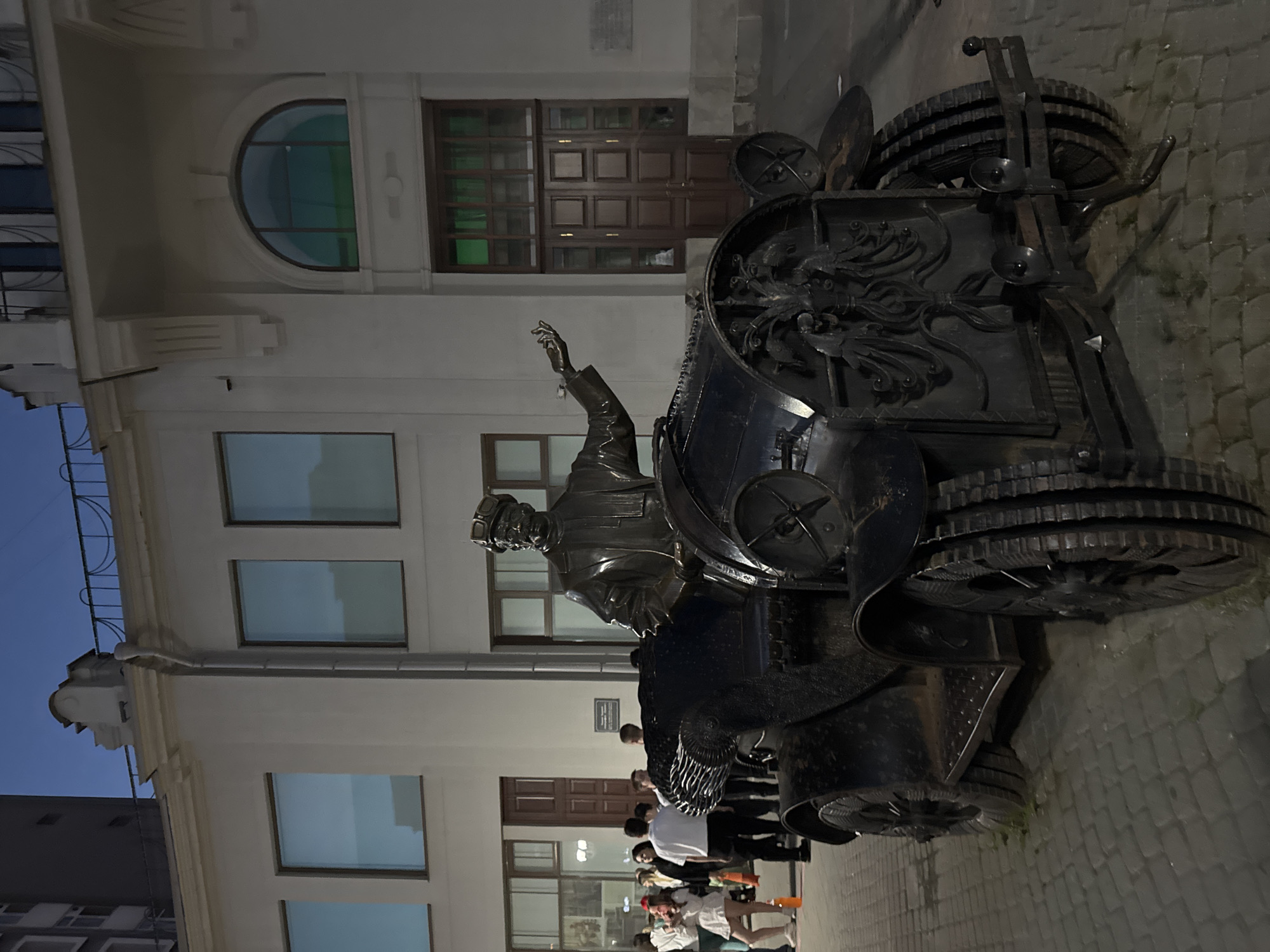 The image shows a statue of a man in a car, located in front of a building. The statue is made of dark metal and depicts a man wearing a hat and goggles, sitting in an old-fashioned car. The car has a decorative front grill. Several people are standing in the background, some looking towards the statue and others walking away. The building behind appears to be an office building or a historical structure, with large windows and architectural details. The scene is set on a cobblestone street, and the sky is a deep blue, suggesting it is dusk or early evening. This is the monument to Gorky and the car "GAZ-A" on the embankment of the Iset River in Yekaterinburg, Russia.