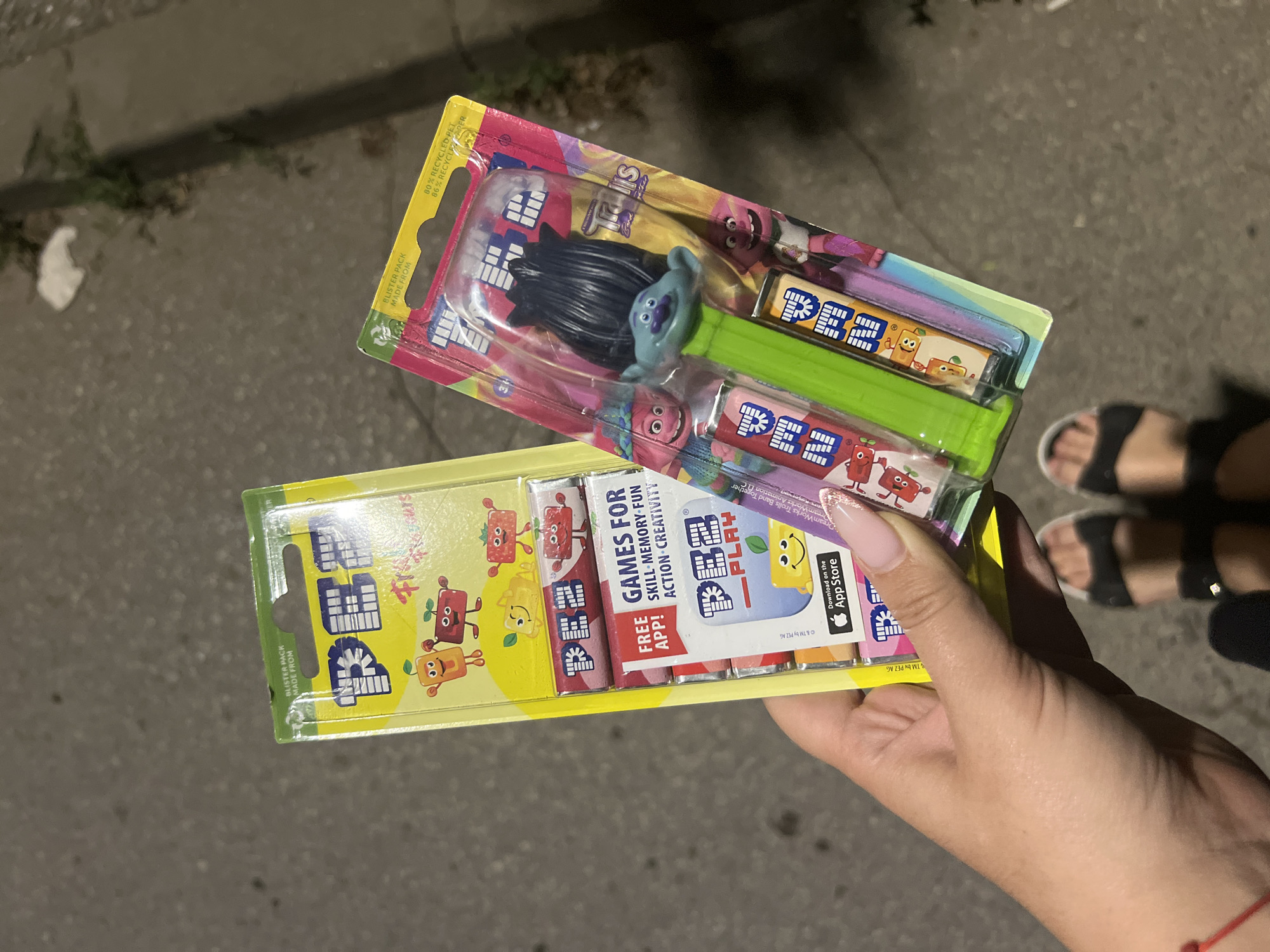 A person's hand holds two blister-packaged PEZ dispensers in an outdoor, paved area, likely a sidewalk or street. The dispenser on the left is yellow and features cartoon fruit characters. It contains three PEZ candy rolls. The dispenser on the right is pink and features characters from the movie "Trolls," specifically a green-headed troll figure atop the dispenser. This dispenser also contains three PEZ candy rolls. The background shows a blurred, dimly lit, textured surface, consistent with asphalt or concrete, and some foliage and debris. The lighting suggests it is nighttime or dusk. The person's feet, wearing black sandals, are visible at the bottom of the frame. The image does not provide any information about the specific location within Sofia, Bulgaria.