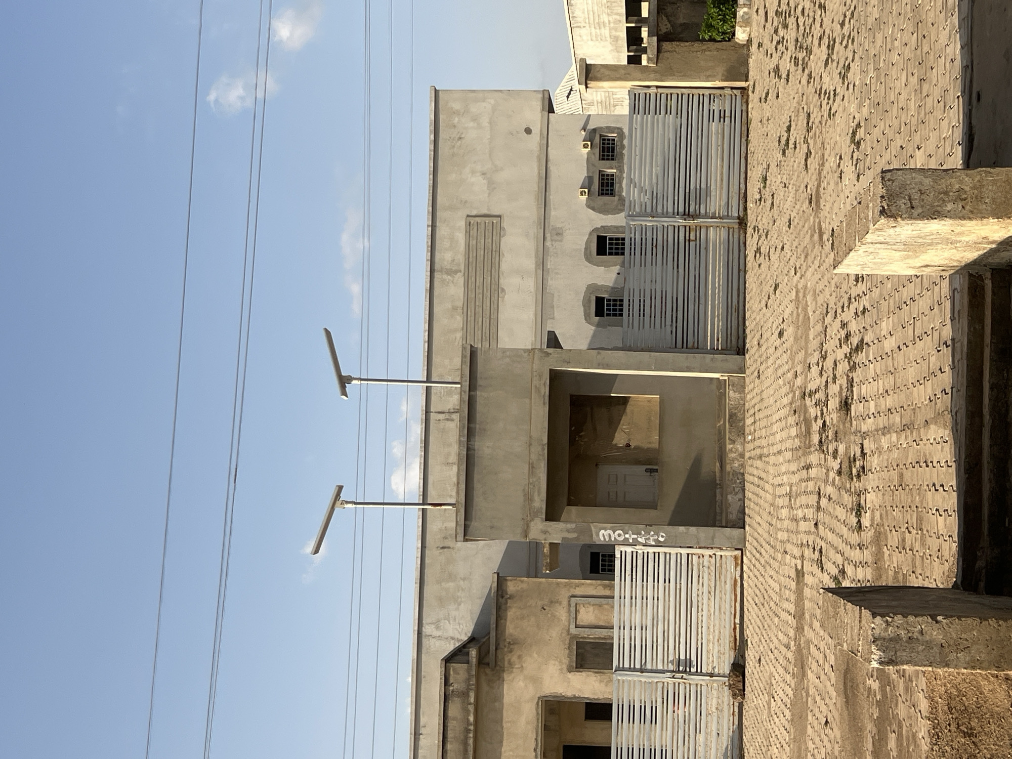 The image displays an exterior street view in the city of Bauchi, Nigeria, featuring a row of unpainted concrete buildings under a clear blue sky with sparse clouds. The upper portion of the frame shows multiple power lines traversing horizontally. Two tall, grey solar-powered streetlights are prominently positioned in front of the buildings.
On the left, a single-story structure presents a weathered concrete facade with open rectangular spaces where windows or doors would be, and is fronted by a horizontal-slat metal gate. Adjacent to it, a central concrete archway or entrance structure partially obscures a white door within. On the right face of this central structure, a vertical inscription reads "304446." The building on the right is a two-story or tall single-story structure with a flat roof, featuring a smooth, unpainted concrete exterior. It includes multiple small, barred windows and horizontal concrete relief detailing on its upper wall, and is also secured by a horizontal-slat metal gate.
The foreground consists of a vast expanse of light-colored interlocking pavers, arranged in a wavy pattern and showing scattered patches of green vegetation. Two rough-hewn concrete blocks, possibly remnants of a low wall or steps, are visible in the immediate foreground. No people, vehicles, or animals are present in the scene. The lighting suggests either early morning or late afternoon, with shadows cast on the ground.