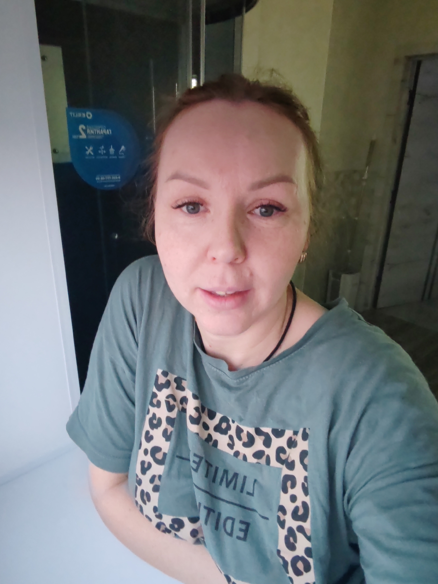 A female individual, appearing to be in her 30s-40s, with light-colored hair pulled back and blue eyes, is depicted from the chest up, facing the camera directly. She wears a dark teal short-sleeved t-shirt featuring a large beige and black leopard print design with vertically aligned, mirrored text reading "ЭТИМИЛ" (LIMITLESS) and "НОЙШИДЕ" (EDITION). A thin black cord necklace is visible around her neck, and a small gold-colored hoop earring is present in her visible left ear.
The subject is situated indoors. To her immediate left, a white vertical surface is present. Adjacent to this and behind the subject's left side is a glass partition or door, reflecting light. Adhered to this glass is a blue oval sticker with white, mirrored text. Prominent mirrored text reads "RNTHANAT," which when reversed correctly identifies "TANHARTN." Additional mirrored text, "TJURQ," is also visible. Below this text are icons depicting a fork, knife, spoon, and a stylized human figure, along with a Russian phone number: "89-22-386-30-60". To the subject's right, the background consists of a light-colored, possibly textured or stuccoed wall, with a partially visible open doorway leading into an adjacent room.
Based on the visible phone number and the mirrored business name "TANHARTN" on the sticker, the precise location is identified as the Cafe-Bar "TANHARTN" in Chaikovsky, Perm Krai, Russia.