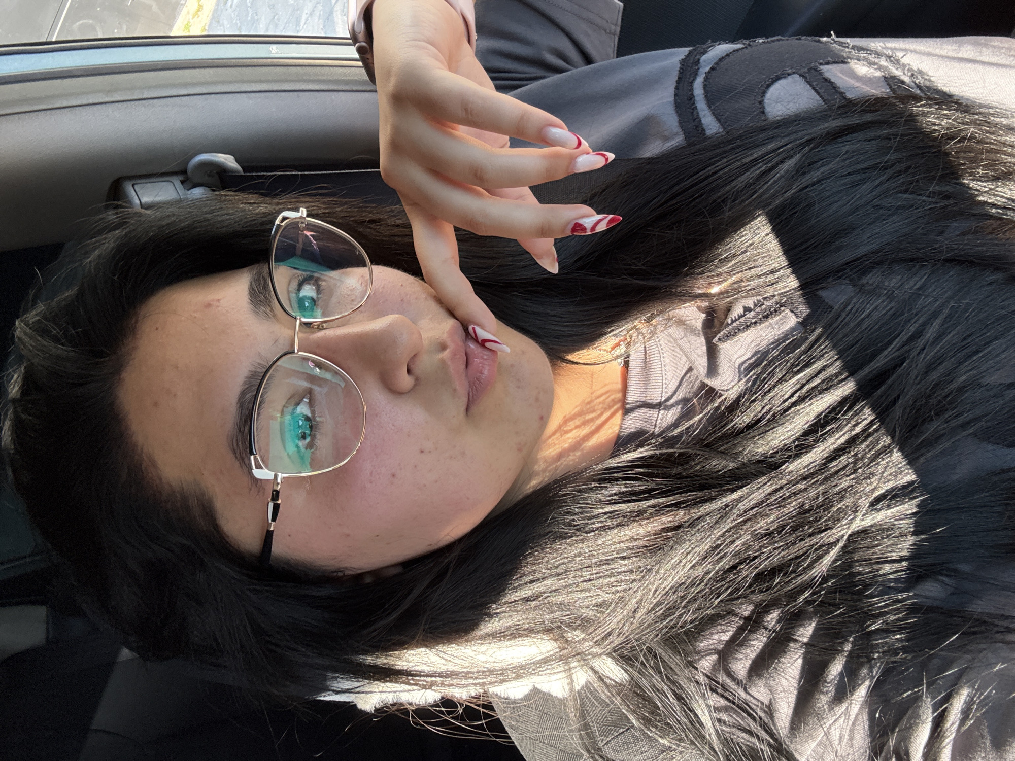 A young woman lies on her side, likely inside a car, wearing glasses and posing with her hand near her face. The woman has black hair and fair skin, with noticeable freckles and some skin imperfections. She is wearing glasses with thin gold frames. Her fingernails are long and painted with a white and red design. The interior of the vehicle is visible. The location and city are not discernible.