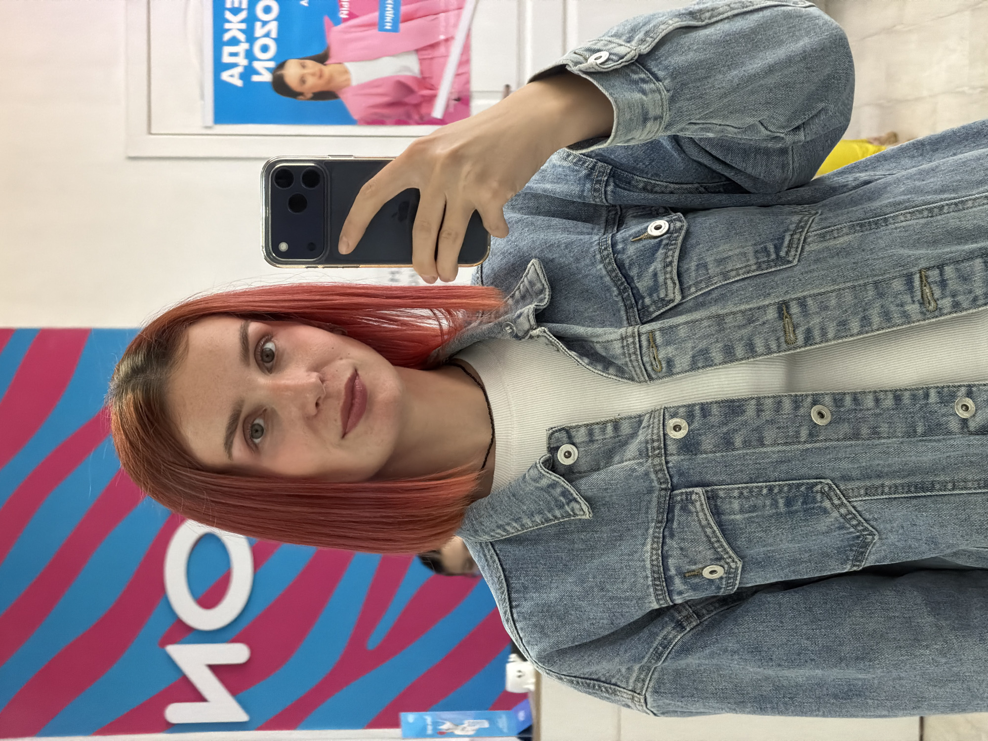 A woman is taking a selfie in what appears to be an indoor commercial space. She holds a dark-colored smartphone in her right hand. She has red hair, is wearing a white top, and a blue denim jacket. Behind her, a wall is decorated with blue and pink stripes and the white Cyrillic letters "M O". A poster with a woman wearing pink is mounted on the wall in the upper background. The location is Chaikovsky, Russia.