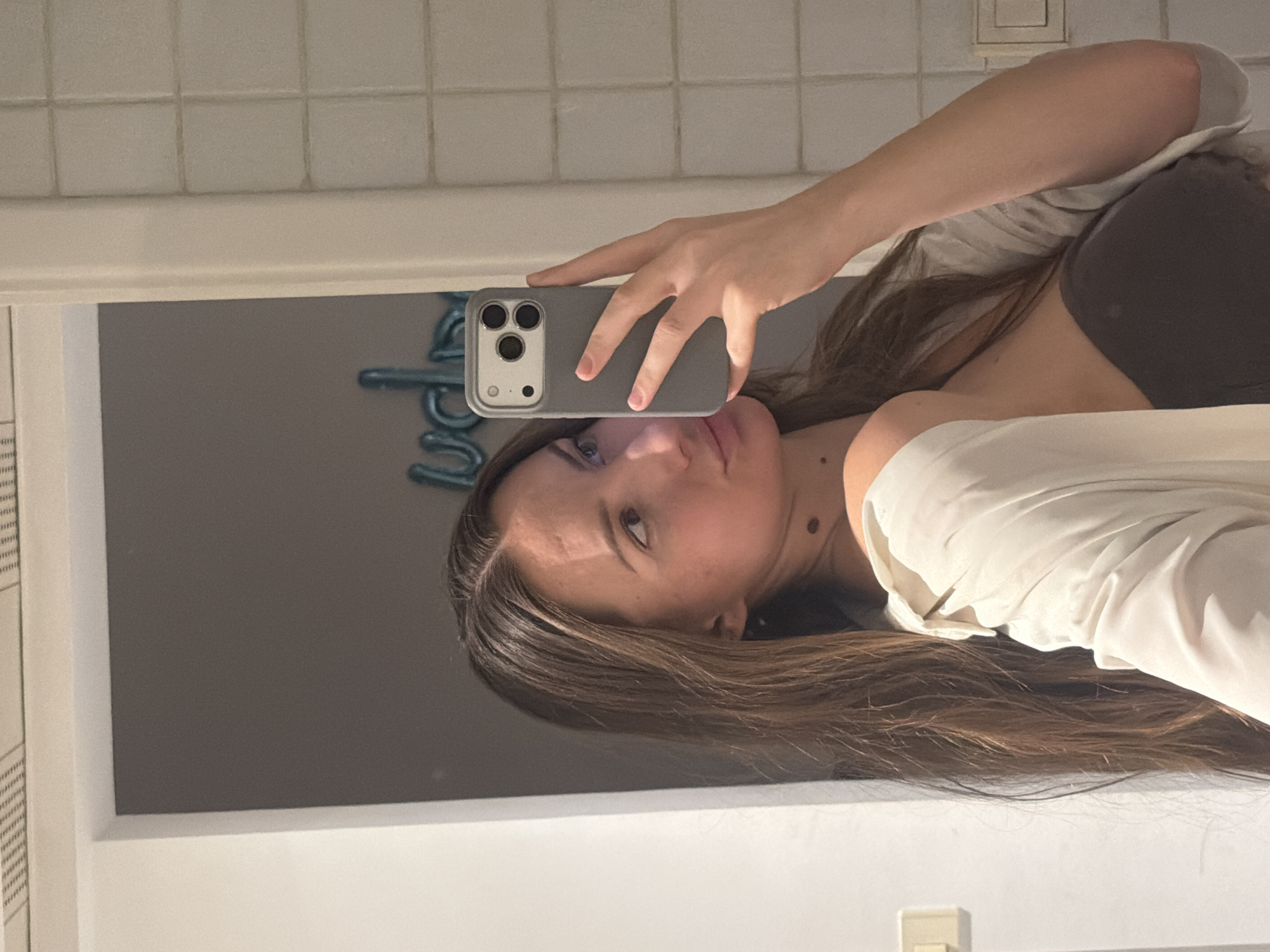 A young woman is taking a mirror selfie in a bathroom. She has long brown hair and is wearing a white shirt over a dark strapless top. She is holding a gray smartphone in her right hand, which is angled to capture her reflection. In the background, there are blue balloons that appear to spell out "LOVE" and a tiled wall. The lighting suggests it is indoors, possibly in a hotel room. The context provided suggests the location is Sharm El Sheikh, Egypt.