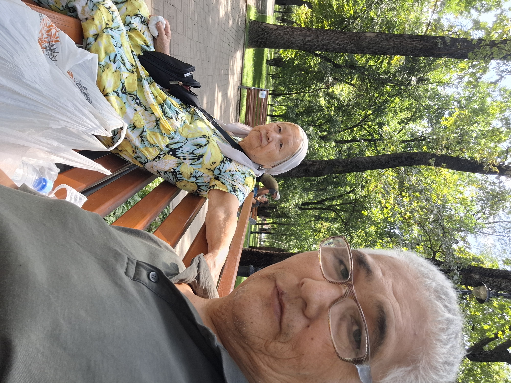Two individuals are seated on a park bench in Almaty, Kazakhstan. The person in the foreground, on the right, is facing the camera and appears to be an adult male with gray hair and glasses. They are wearing a dark gray polo shirt. To the left, an elderly woman is also seated on the bench, looking towards the camera. She has gray hair and is wearing a headscarf and a yellow and blue floral dress. In front of the woman are two white plastic shopping bags. The bench is made of wood and is situated in a park with trees and grass. The background shows more trees and a pathway. The lighting suggests it is daytime, likely sunny.