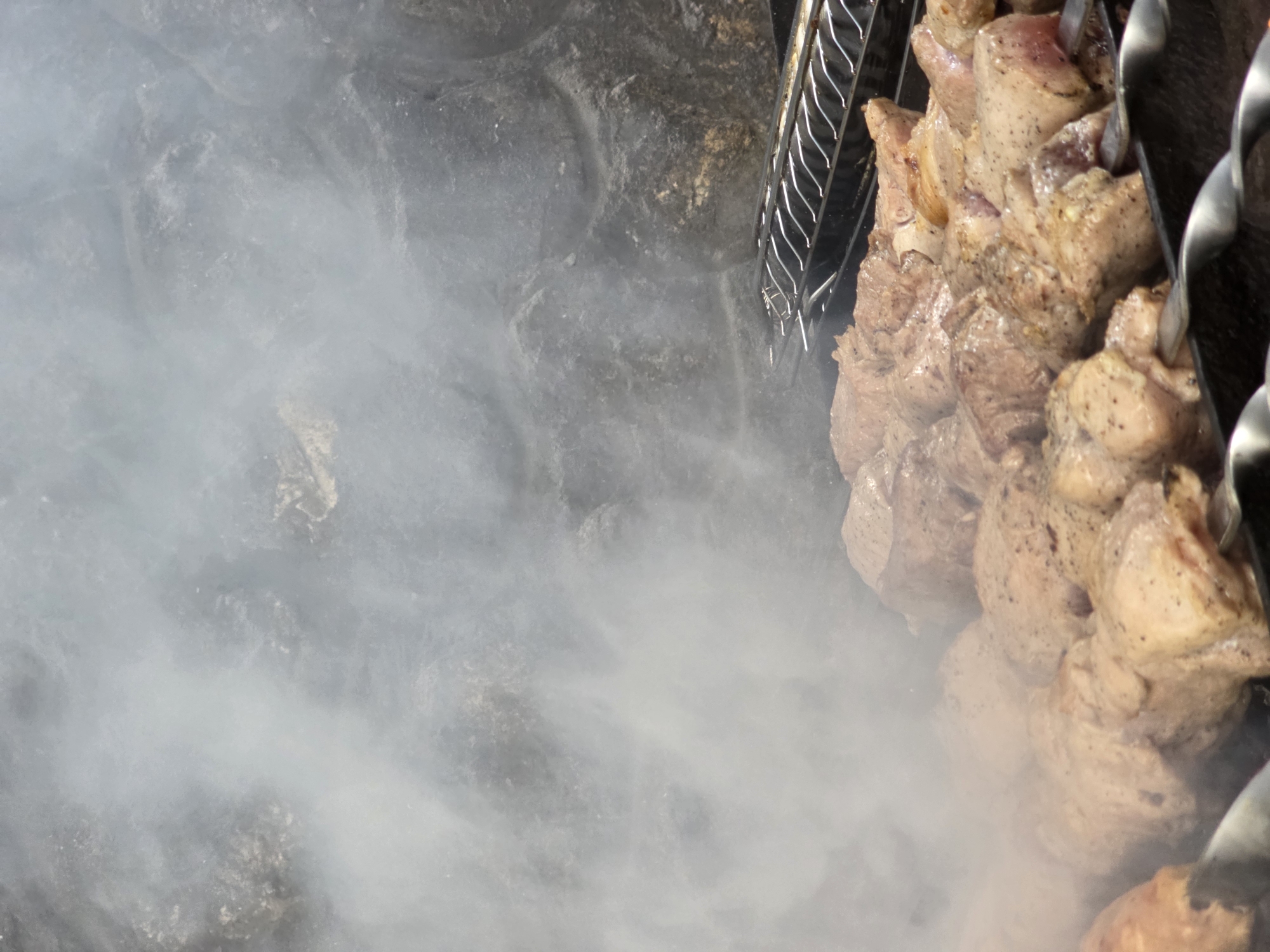 In Pidhorodne, Ukraine, skewers of marinated meat are being grilled over a fire, producing a significant amount of smoke that obscures the background. The meat, cut into uniform pieces, is skewered on metal rods and arranged on a grill. The grill itself is placed in front of a rough, stone-like surface, possibly a wall or an outdoor oven. A second grill grate is visible in the upper right portion of the image. The focus is on the meat and the smoke, suggesting an outdoor cooking event.