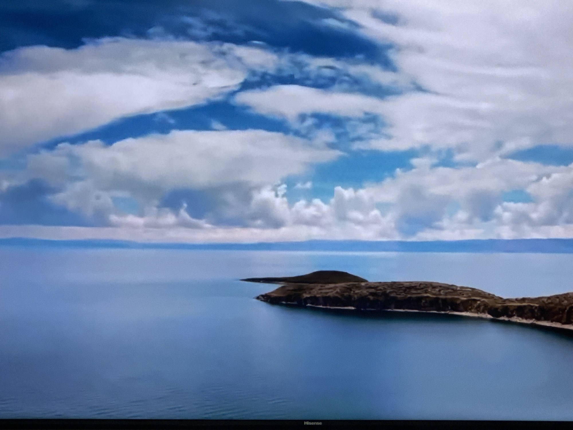 The image depicts a serene aerial view of islands in a vast body of water under a dramatic sky. The water is a deep, tranquil blue, with subtle ripples visible. Two distinct islands, characterized by their brown, textured terrain, emerge from the water. The larger island forms a crescent shape, while a smaller, conical island is situated to its left. The horizon line is a thin, hazy band separating the water from a distant landmass. Above, the sky is a vibrant spectacle of blue interspersed with voluminous white clouds, some of which are tinged with gray, suggesting depth and movement. The scene is devoid of any human presence. The branding "Hisense" is visible at the bottom edge of the image, indicating it is a photograph of a television screen. There is no information available to suggest this scene is located in the city of Ləcət, Azerbaijan.
