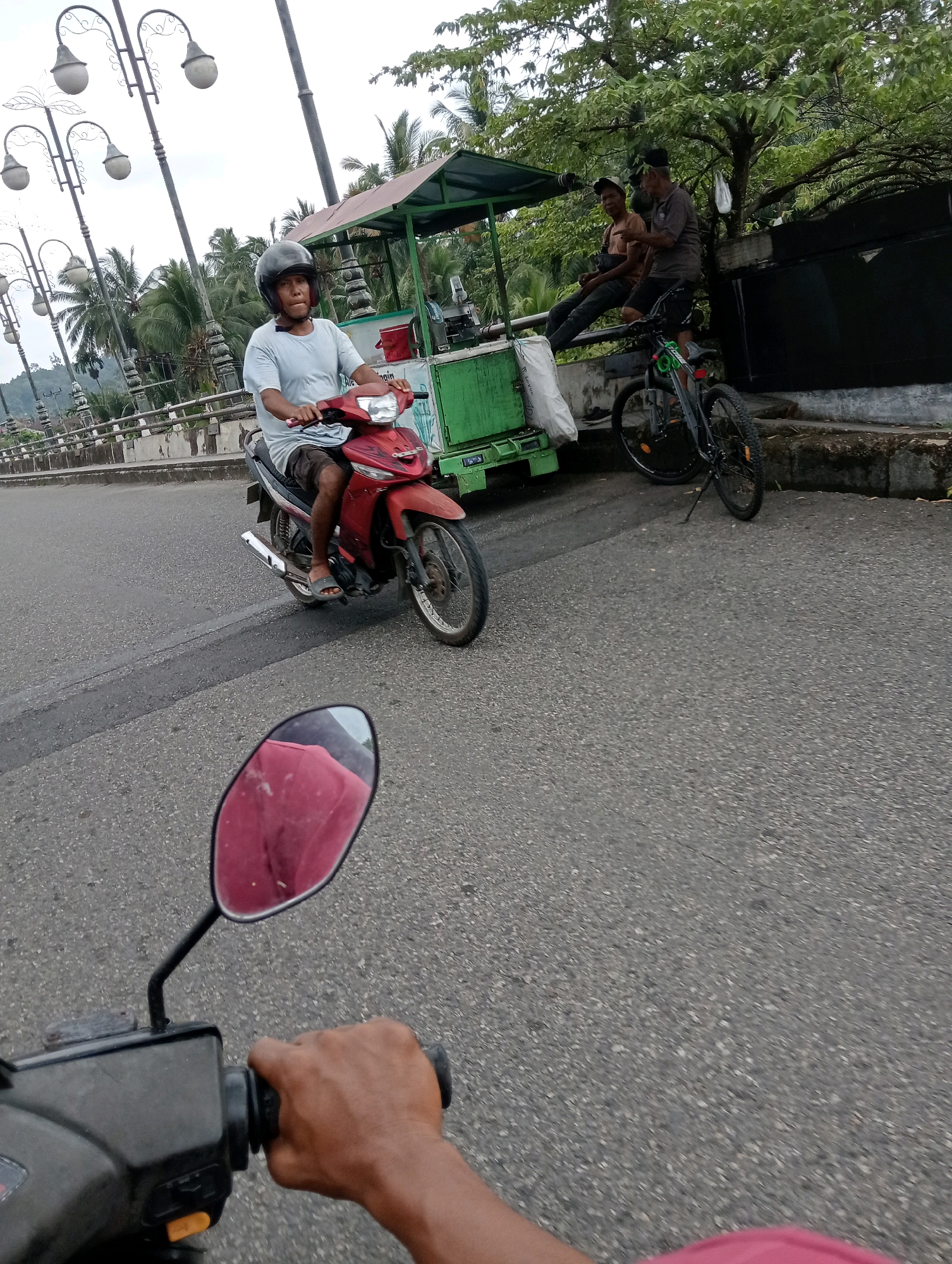 In Campago, Indonesia, a motorcyclist with a black helmet rides a red motorcycle on a paved road. The rider wears a light blue t-shirt and dark shorts. Behind the motorcyclist, two men are seated on a concrete barrier next to a green food cart. One man wears a black cap and a tan t-shirt, while the other wears a dark t-shirt and shorts. A bicycle is parked next to the food cart. Palm trees and other foliage form the background. The image is taken from the perspective of another motorcyclist, with the handlebar and a rearview mirror visible in the foreground.