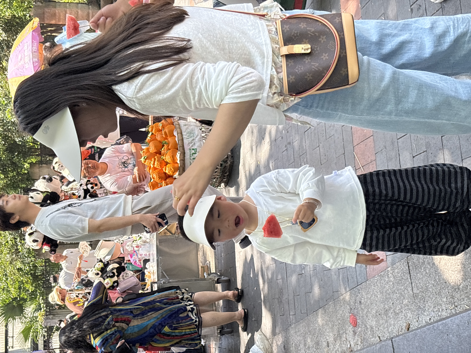 On a paved pedestrian street in Jiulongpo District, China, a woman wearing a white shirt, blue jeans, and a brown handbag adjusts a white visor on a child wearing a white long-sleeved shirt and black and gray striped pants. The child is holding a piece of watermelon on a stick. A man wearing a white t-shirt holds a phone. Vendors display goods, including stuffed pandas and oranges in woven baskets. Another woman stands nearby wearing a blue and gold patterned dress.