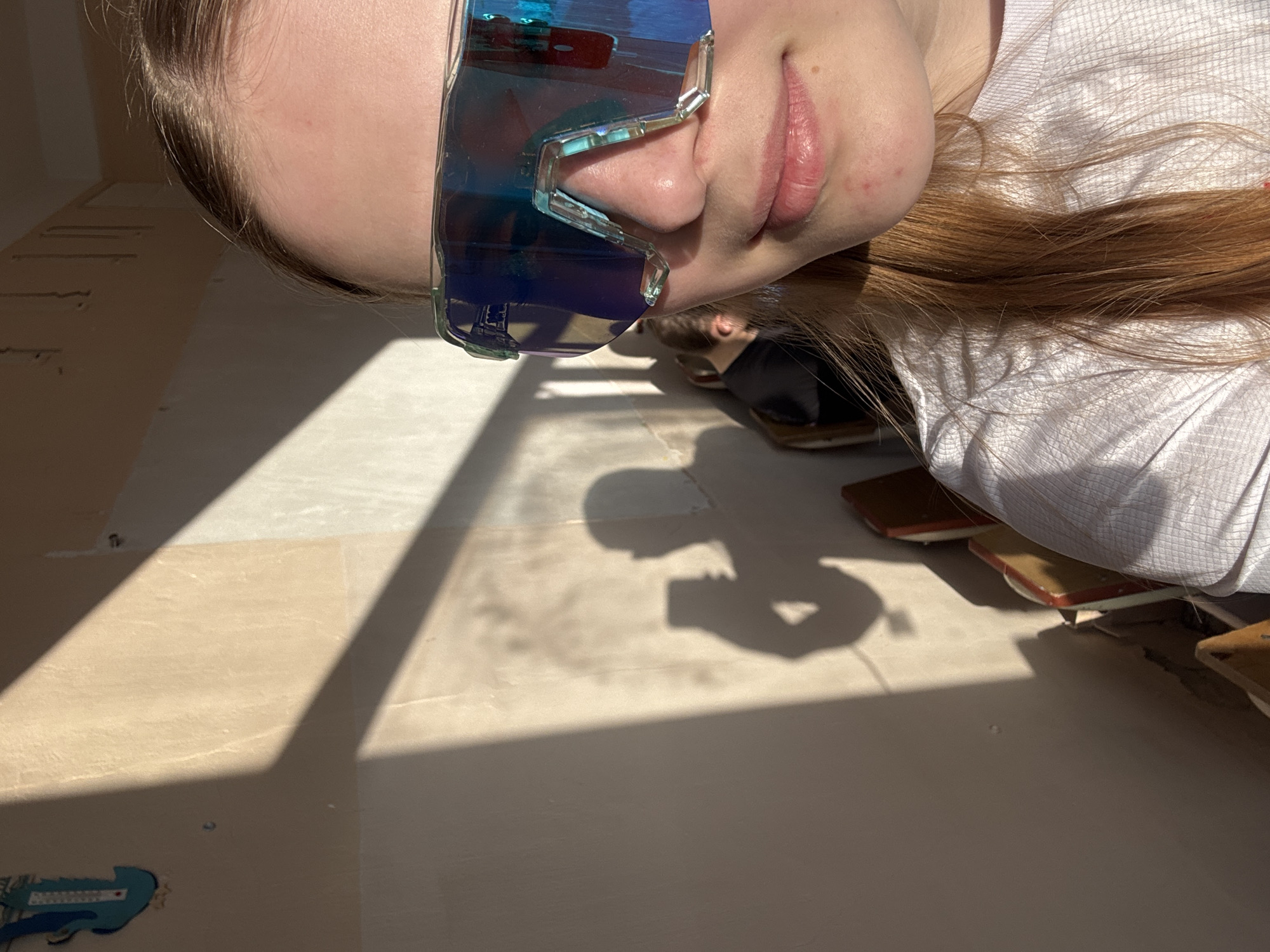 A person wearing blue mirrored sunglasses is lying on a tan-colored surface, possibly a floor, that is undergoing renovation. The floor has sections of exposed subfloor and some trim pieces scattered around. The person's shadow is cast on the floor, and part of another person is visible in the background. A blue tool or item of equipment is in the lower left corner. The scene is lit by sunlight. There is no information in the image to suggest this is in Usolye, Russia.