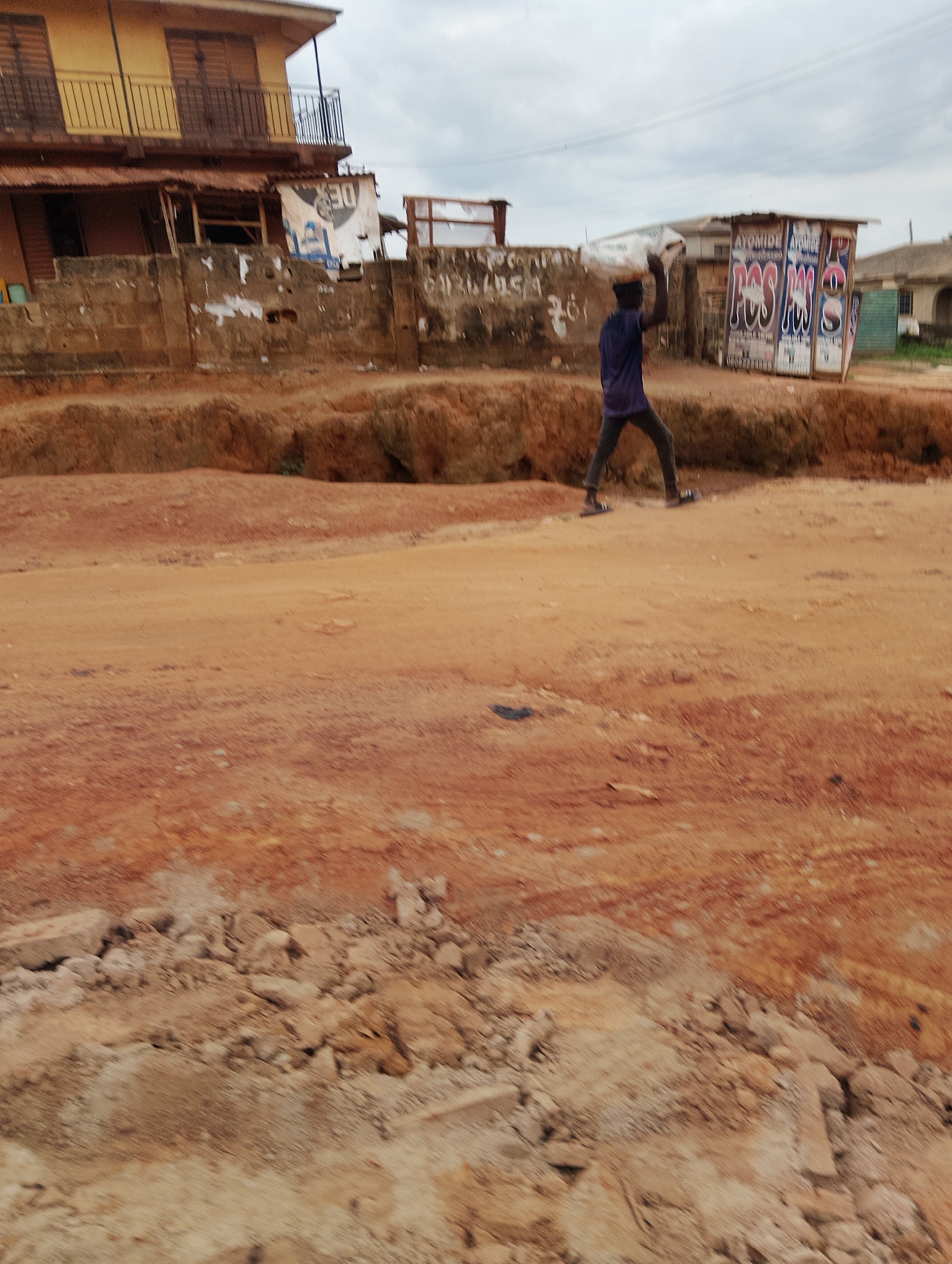 An undated, eye-level, full shot captures a person carrying a load on their head on a dirt road in front of some buildings in Agege, Nigeria. The subject, wearing a blue shirt and dark pants, walks from left to right across the frame. To the left of the subject is a yellowish two-story building with balconies. In front of the building is a wall with posters plastered on it. To the right of the subject is a kiosk with advertisements on its sides. The ground is composed of red dirt and rocks. The sky is overcast.