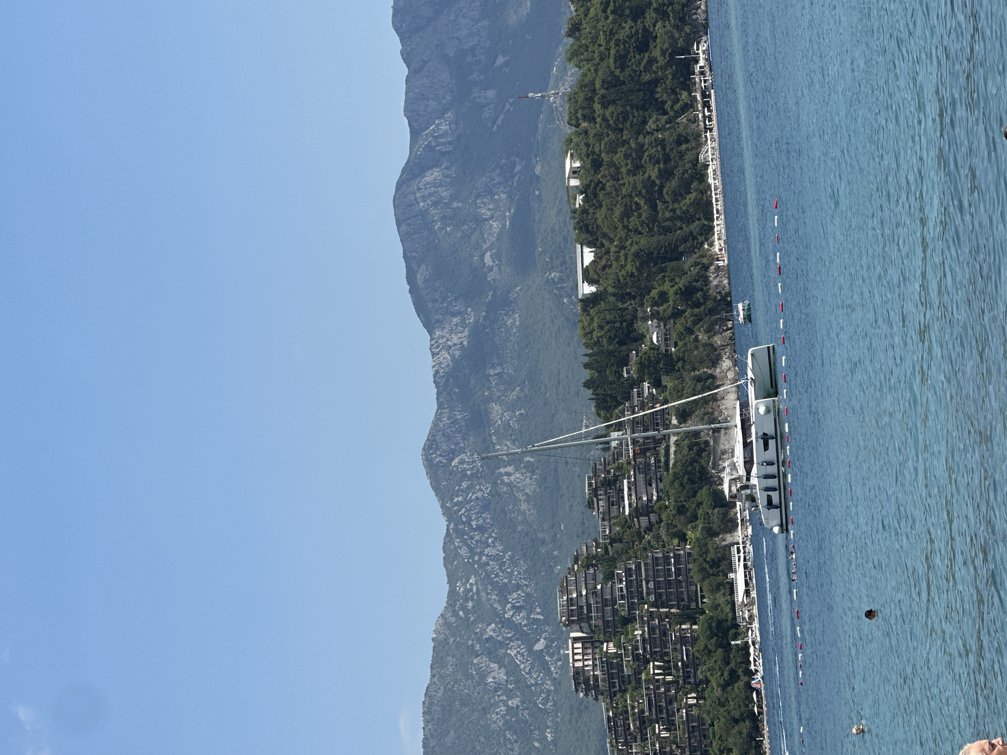 A wide shot captures a serene day at the beach in Budva, Montenegro, with the crystal-clear turquoise waters of the Adriatic Sea dominating the foreground. A sleek white catamaran, its sails furled, floats in the middle distance, a testament to the area's popular leisure activities. A line of buoys marks a swimming area, with several people visible enjoying the water, some swimming and others wading near the shore.
In the background, the coastline is dotted with lush green trees, behind which a complex of multi-tiered buildings with cascading balconies, characteristic of modern resort architecture, is nestled into the hillside. Towering above the resort and greenery are the rugged, rocky slopes of a mountain range, their peaks disappearing into the clear blue sky. A single, slender telecommunications tower stands out against the mountainous backdrop. The scene is bathed in bright sunlight, indicating a warm, summer day.
