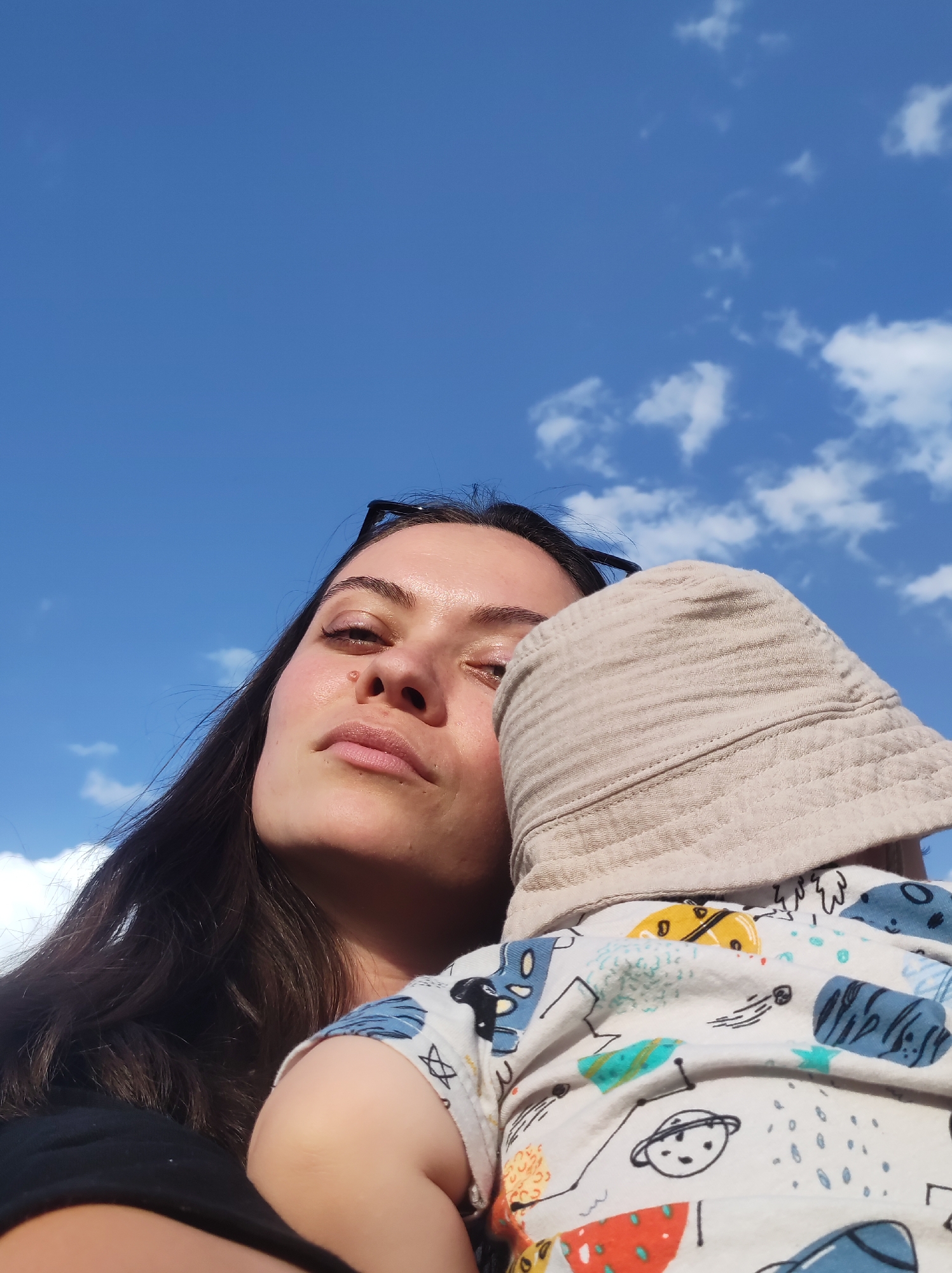 A low-angle medium shot depicts a fair-skinned woman with long, dark hair holding a baby. The woman gazes upwards, her expression neutral, while the baby is partially obscured by a light beige sun hat. The baby wears a white patterned shirt featuring illustrations of planets, stars, and abstract shapes. The background consists of a blue sky dotted with fluffy white clouds. The location, specified as Chaikovsky, Russia, is not visually evident in the image.