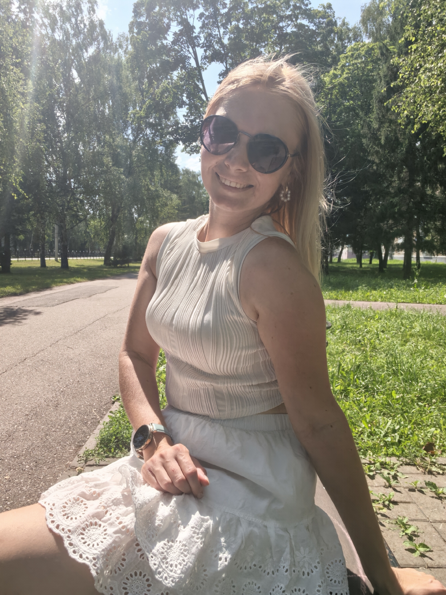 A blonde woman with sunglasses smiles at the camera, sitting outdoors on a sunny day. She is wearing a white, sleeveless ribbed top and a white lace skirt. A watch with a light blue band is visible on her left wrist. She is seated on a stone bench next to a paved path, with green grass and trees in the background. The location appears to be a park in Sterlitamak, Russia.