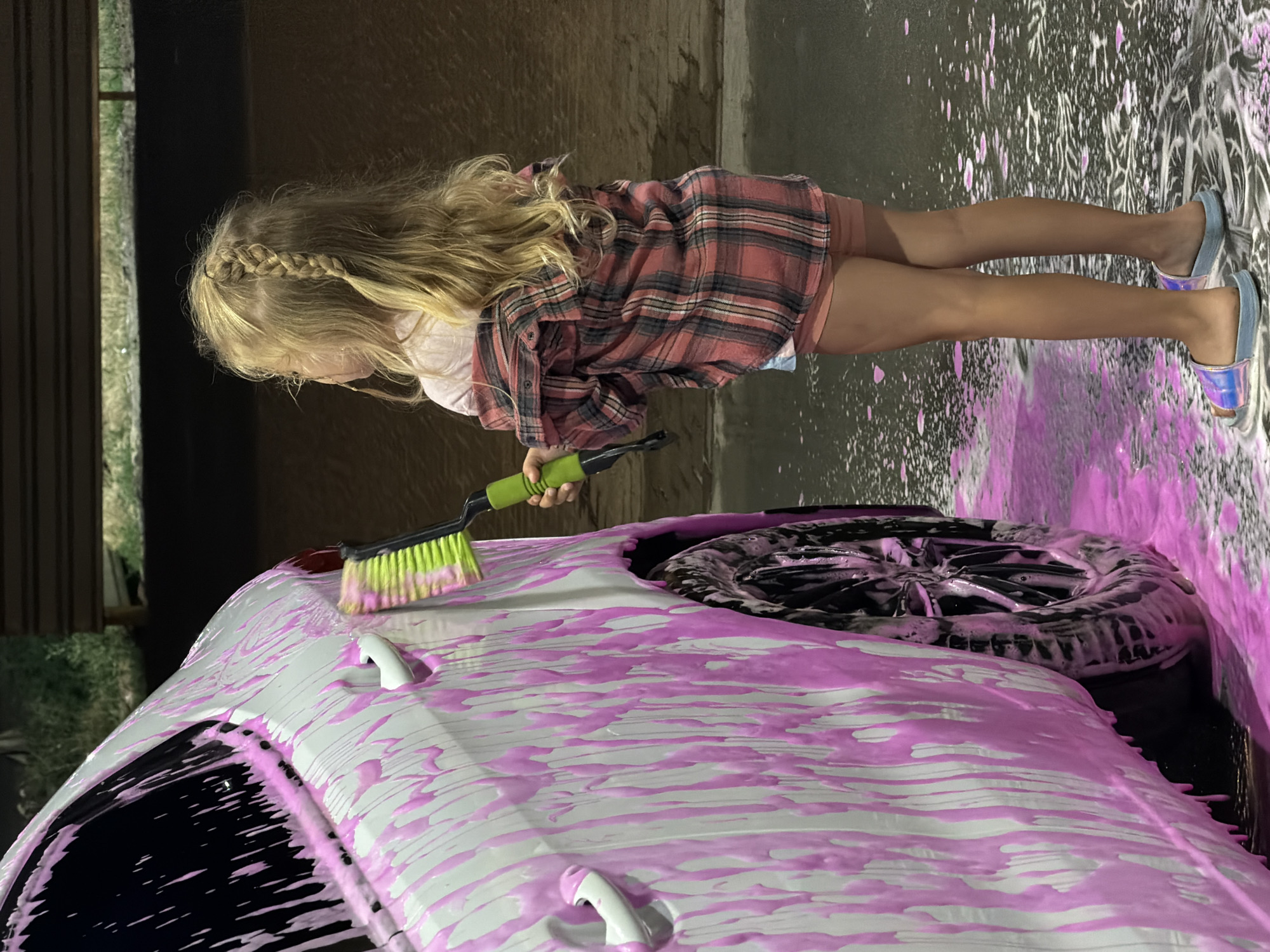 A young girl with long, blonde hair braided down one side is washing a white car. She is wearing a pink and black plaid shirt, pink shorts, and iridescent slides. She is holding a green and yellow brush and is in the process of scrubbing the car's hood, which is covered in pink foam. The car also has pink foam on its windshield and on the ground around it, indicating it is being washed with brightly colored soap. The background is dark, suggesting it might be evening or night. The scene appears to be an outdoor car wash. There is no visible text in the image.