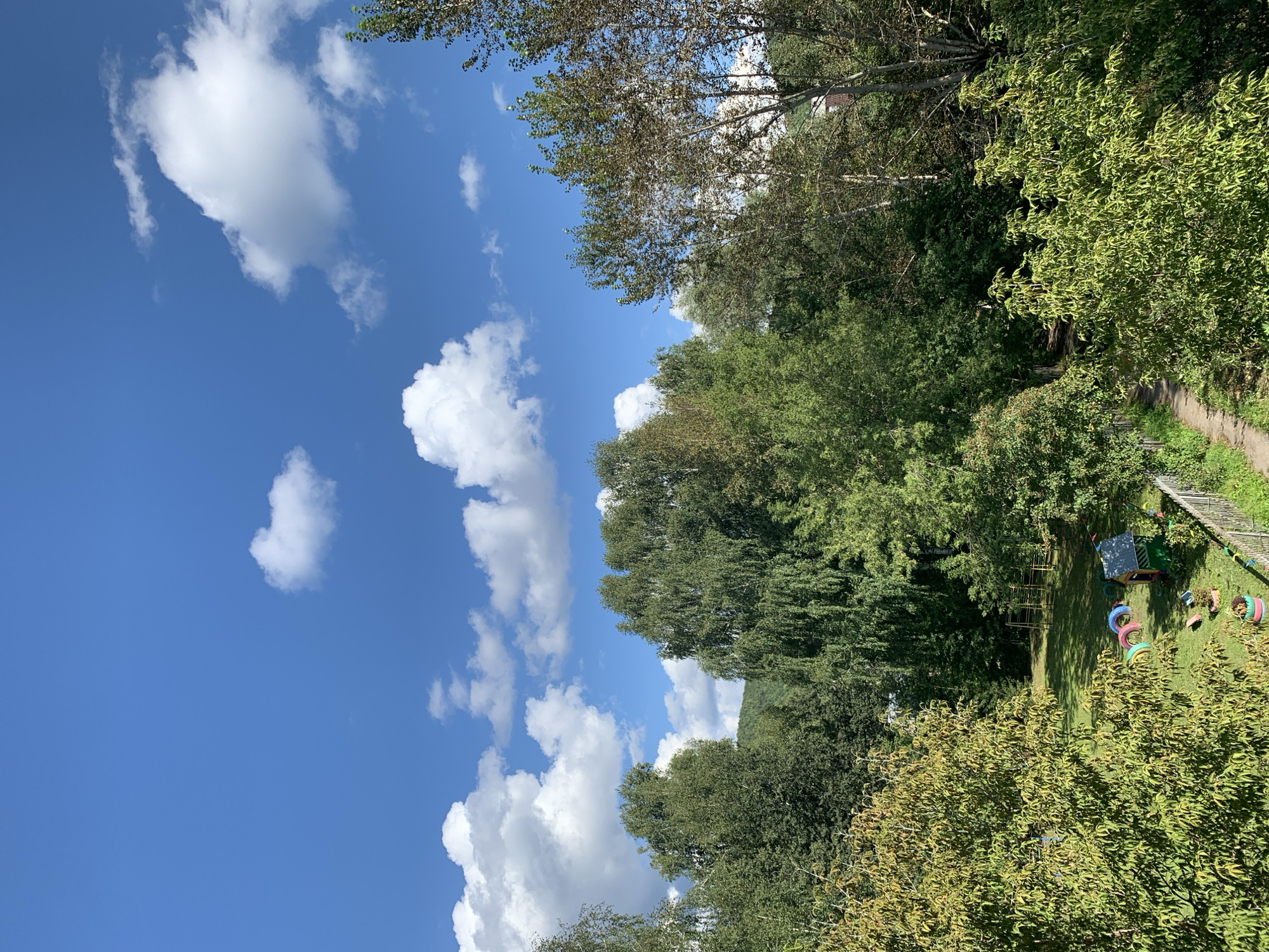 This is a daytime, eye-level, medium shot of a playground in Asha, Russia, framed by lush green trees and a bright blue sky dotted with white cumulus clouds. The playground features a small, colorful playhouse, a set of tire swings, and what appears to be a climbing frame in the background. A dirt path runs along the right side of the playground, bordered by a white wooden fence. The overall impression is one of a serene, sunlit day in a natural, verdant setting.