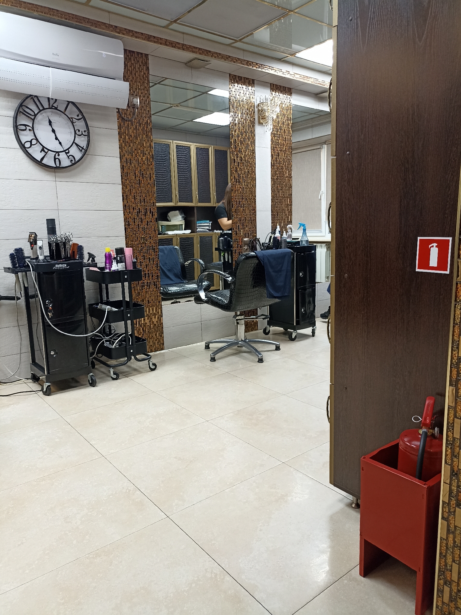 The image depicts the interior of a well-lit hair salon, likely located in the city of Заринск, Russia. The scene is dominated by cream-colored tiled flooring and light-colored wood-paneled walls. To the left, a large circular clock with black Roman numerals hangs on the wall, accompanied by a white air conditioning unit above it.

In the foreground, a black trolley cart is laden with hair styling tools, including brushes, scissors, clippers, and various bottles of hair products. Beside it, another smaller, three-tiered black trolley holds more bottles and equipment. A black salon chair with a textured seat and chrome base is positioned centrally, with a dark blue towel draped over its back.

The background reveals mirrored walls adorned with a decorative mosaic of small, rectangular tiles in shades of brown and gold. Behind the salon chair, several cabinets with glass doors and dark frames are visible, containing what appear to be folded towels and other salon supplies.

To the right of the image, a large, dark brown wooden door is partially open, revealing a glimpse of the outside or another room. Affixed to the door is a red and white square sign with a white icon of a fire extinguisher. Next to the door, a red metal stand holds a red fire extinguisher. A person, whose face is not clearly visible, is seen from the shoulders down on the far right of the image, wearing dark clothing. The overall impression is that of a functional and organized hair salon environment.