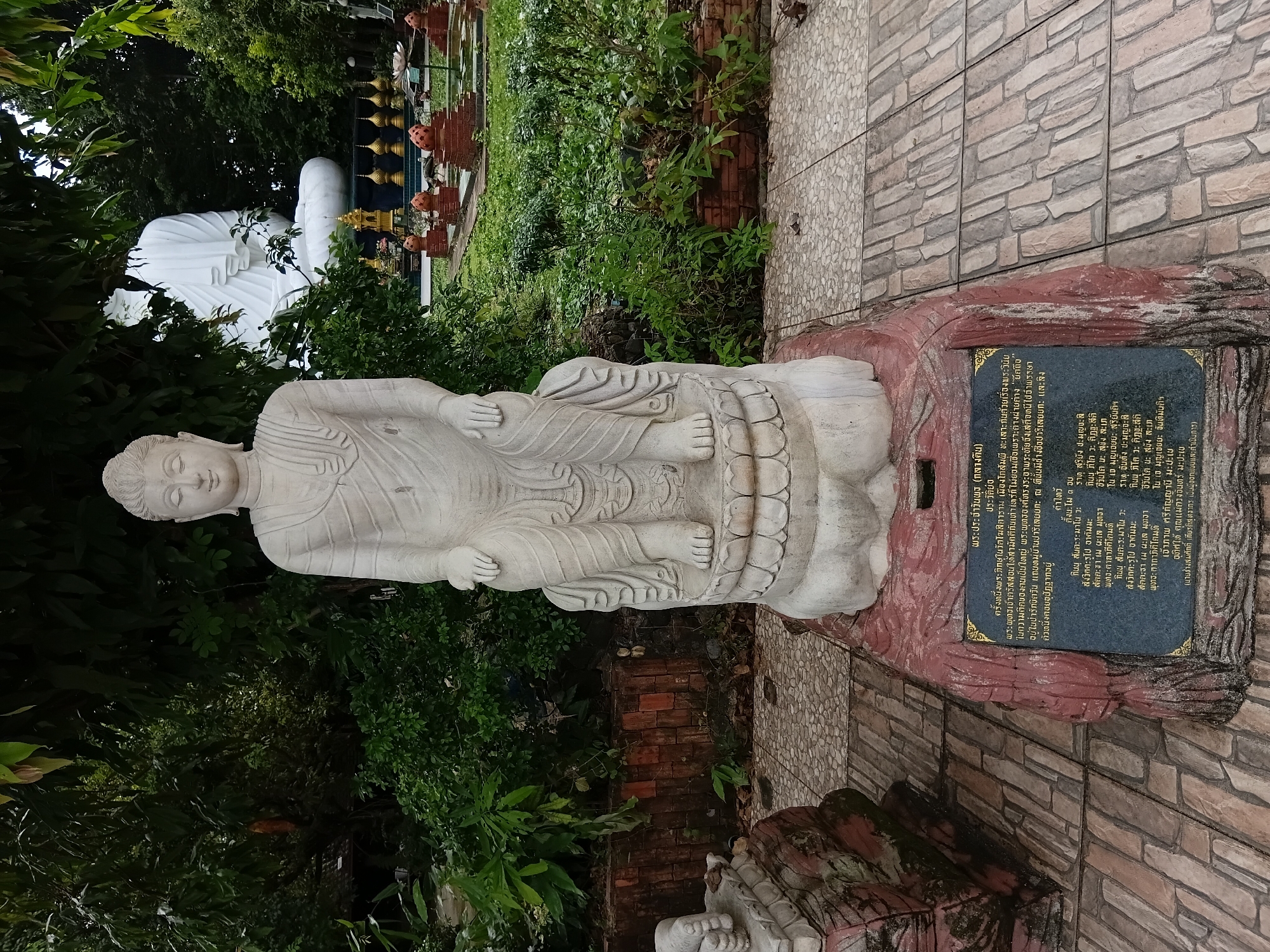 The image shows a stone statue of a meditating Buddha in the city of Ban Ruammit, Thailand. The statue is seated on a lotus pedestal, with its hands resting in its lap. The statue is white and appears to be made of marble or limestone. In the background, there is a larger, white Buddha statue, also seated, surrounded by lush green foliage and other decorative elements typical of a Buddhist temple or garden. A plaque with Thai writing is visible at the base of the foreground Buddha statue. The ground is paved with interlocking stones.