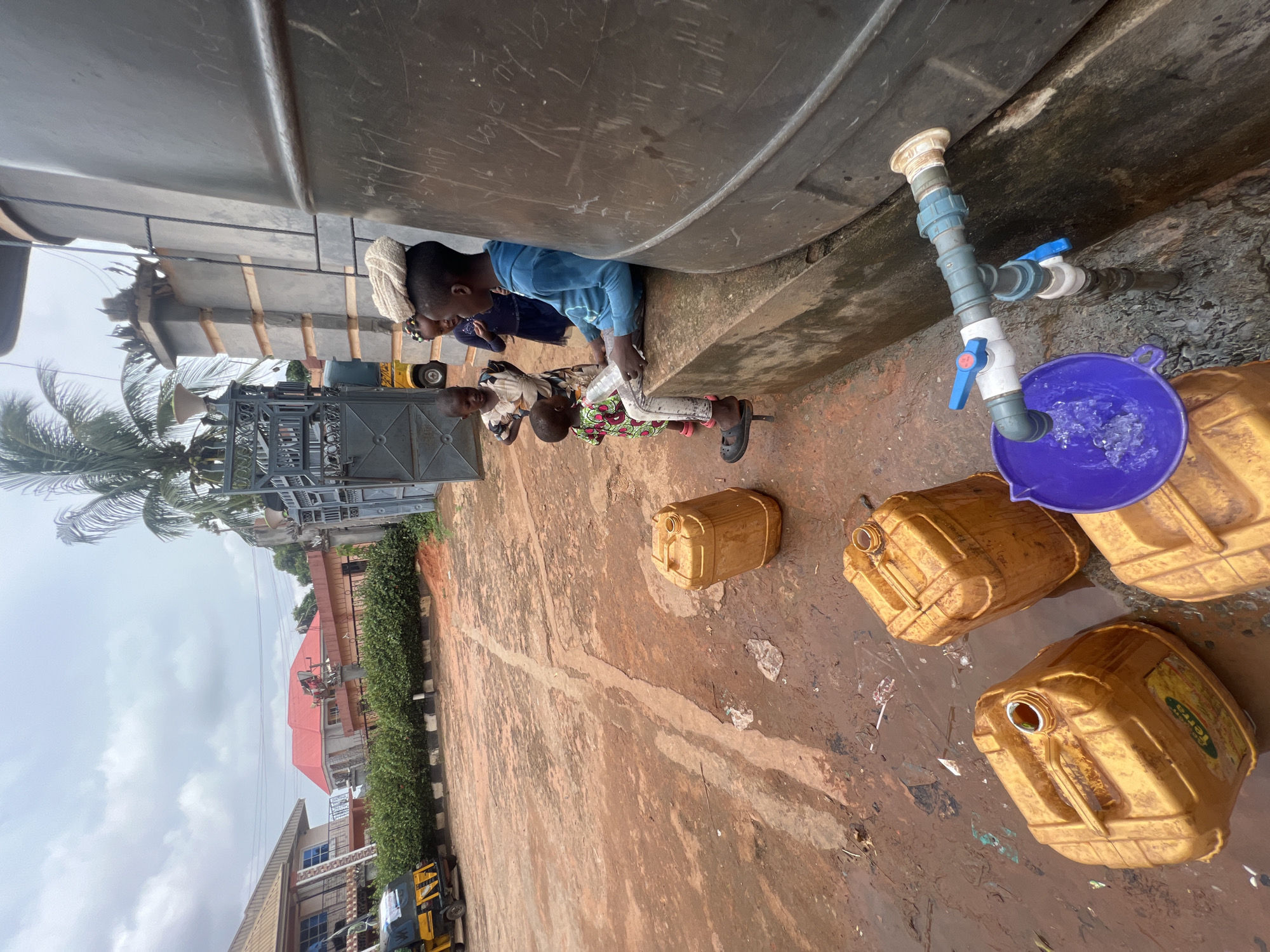 In Obosi, Nigeria, several children are gathered around a large, elevated water tank. They appear to be collecting water from a tap. The ground is muddy. There are several yellow jerrycans nearby, presumably to be filled with water.