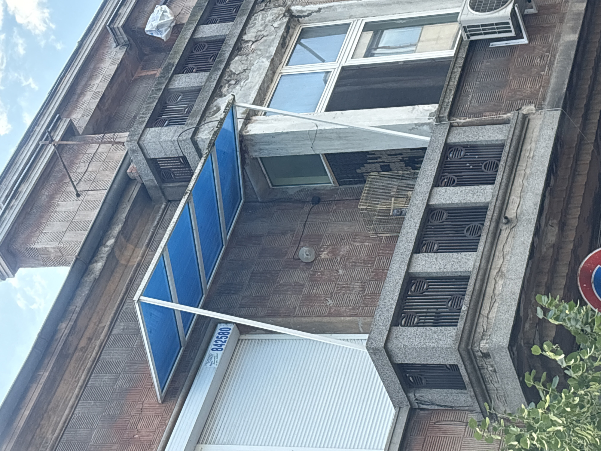 The image shows the exterior of an old apartment building in Burgas, Bulgaria. The building has a facade of reddish-brown brick with decorative elements in grey stone. Several windows are visible, some with blue awnings or sunshades. An air conditioning unit is mounted near one of the windows. There is also a rolled-up metal shutter on a ground-floor window or entrance. The sky is visible in the upper left corner, with some clouds, suggesting it is daytime. A plant with green leaves is visible in the lower right corner. The building appears to be in an urban setting.
Notable details include the texture of the brickwork, which is a repeating pattern of raised lines. The grey stone sections have carved geometric patterns. A small white object wrapped in plastic is attached to the building near the top left. A birdcage is visible on a ledge below a window. A blue and white label with the text "842580" and some other unreadable text is attached to the blue awning. A circular sign with red and blue colors is partially visible on the right edge of the image. The overall impression is one of an aged building in a sunny, possibly warm, climate.