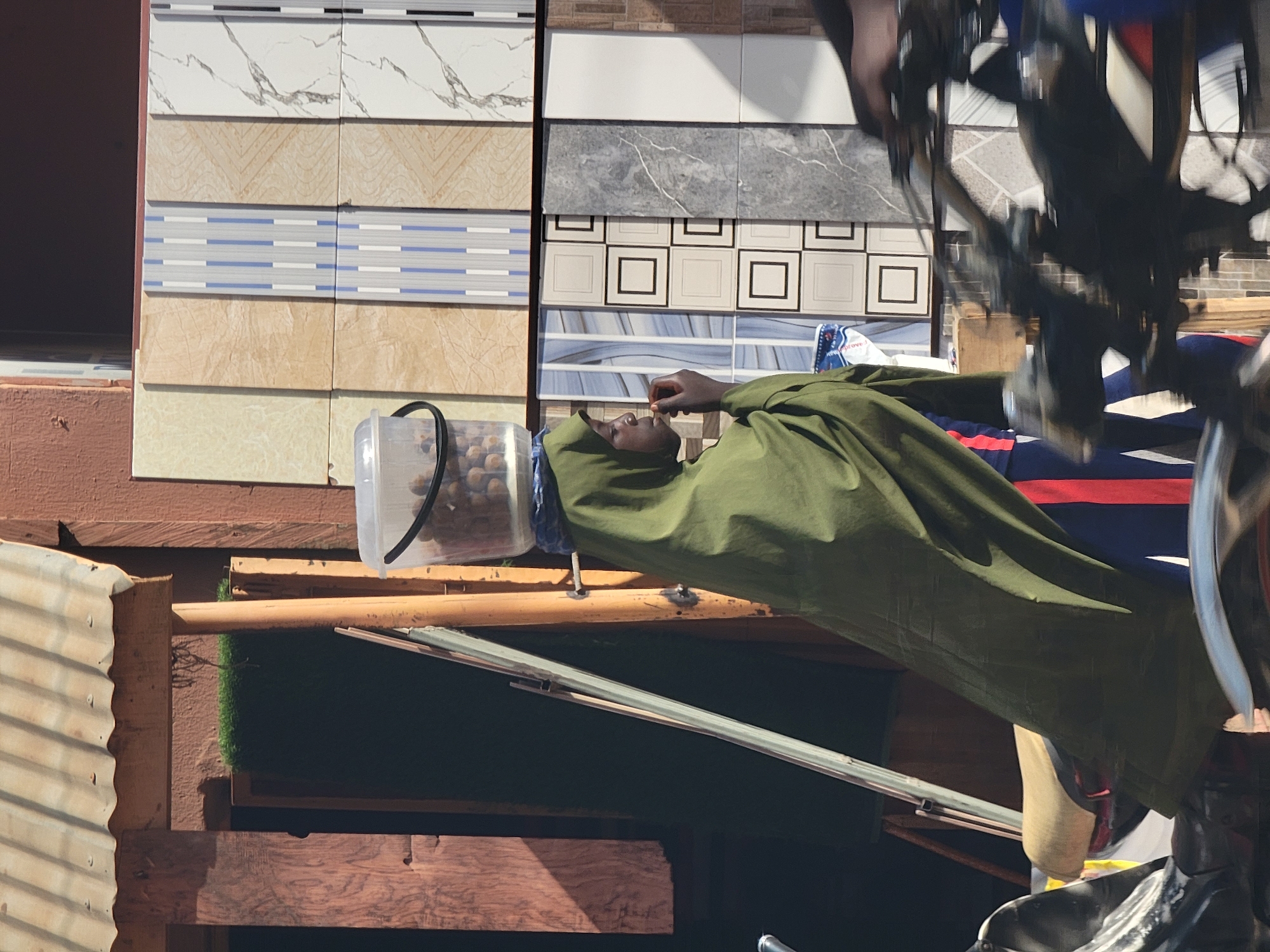 The image shows a person in Maiburiji, Nigeria, dressed in an olive-green garment with a head covering, holding something to their mouth. They are positioned beside a display of ceramic tile samples. A clear plastic container filled with round objects, likely food items, sits nearby. A bicycle is positioned prominently to the right, partially obscuring the view. The background includes corrugated metal, wooden supports, and artificial turf. The scene appears to be an outdoor market or commercial area.
