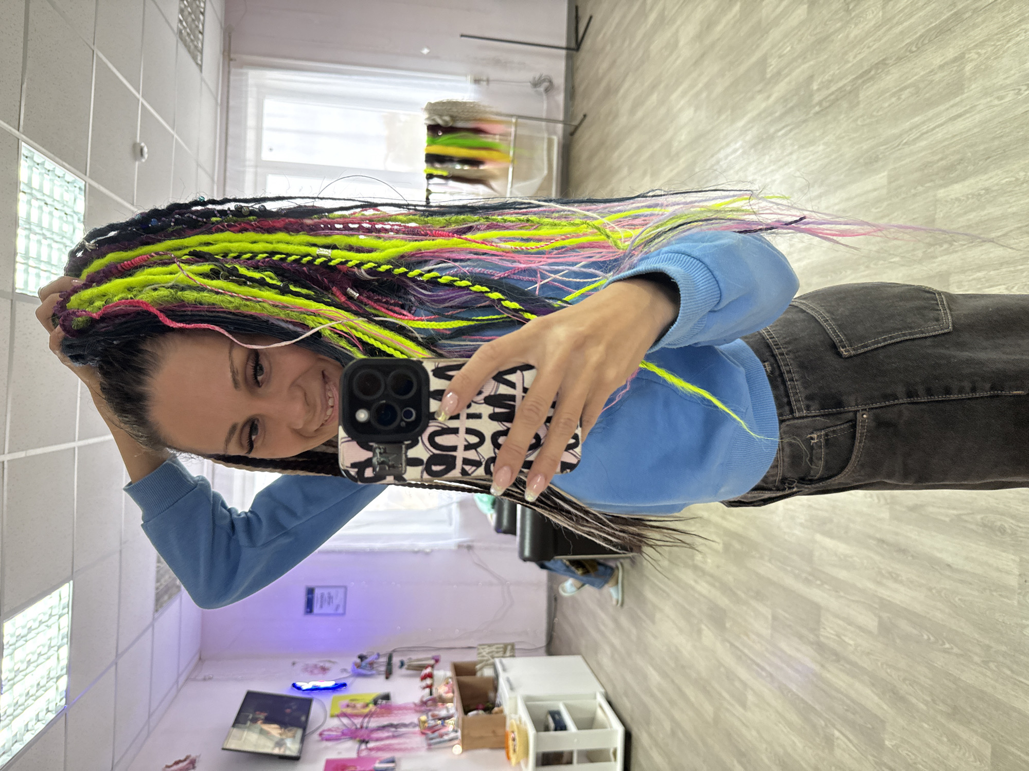A young woman is captured in a mirror selfie, smiling broadly and showcasing her long, vibrant, multicolored dreadlocks or braids. The image is taken indoors, likely in a brightly lit salon or studio in Rusanovo, Russia.
The woman holds a smartphone with a black and white patterned case in her right hand, positioned to capture her reflection. Her hair features a striking combination of black, neon yellow/green, bright pink, and purple strands, some accented with small silver beads or wraps. She is wearing a light blue long-sleeved sweatshirt and dark grey/black jeans. Her manicured fingernails display a light-colored tip.
The background reveals a well-lit room with light-colored wooden or laminate flooring and white tiled walls. A large window in the upper left suggests natural daylight, complemented by indoor fluorescent lighting panels on the ceiling. In the deeper background, a rack displays various colorful hair extensions or braids, indicating a professional hair styling environment. Other objects include a white multi-tiered storage unit with compartments and miscellaneous items on shelves or the floor.
The woman's joyful expression and the prominent display of her intricate, colorful hairstyle suggest she is either celebrating a new look or simply happy with her appearance. The time of day appears to be daytime due to the visible natural light from the window. The phone case features a repeating pattern of stylized letters or symbols.