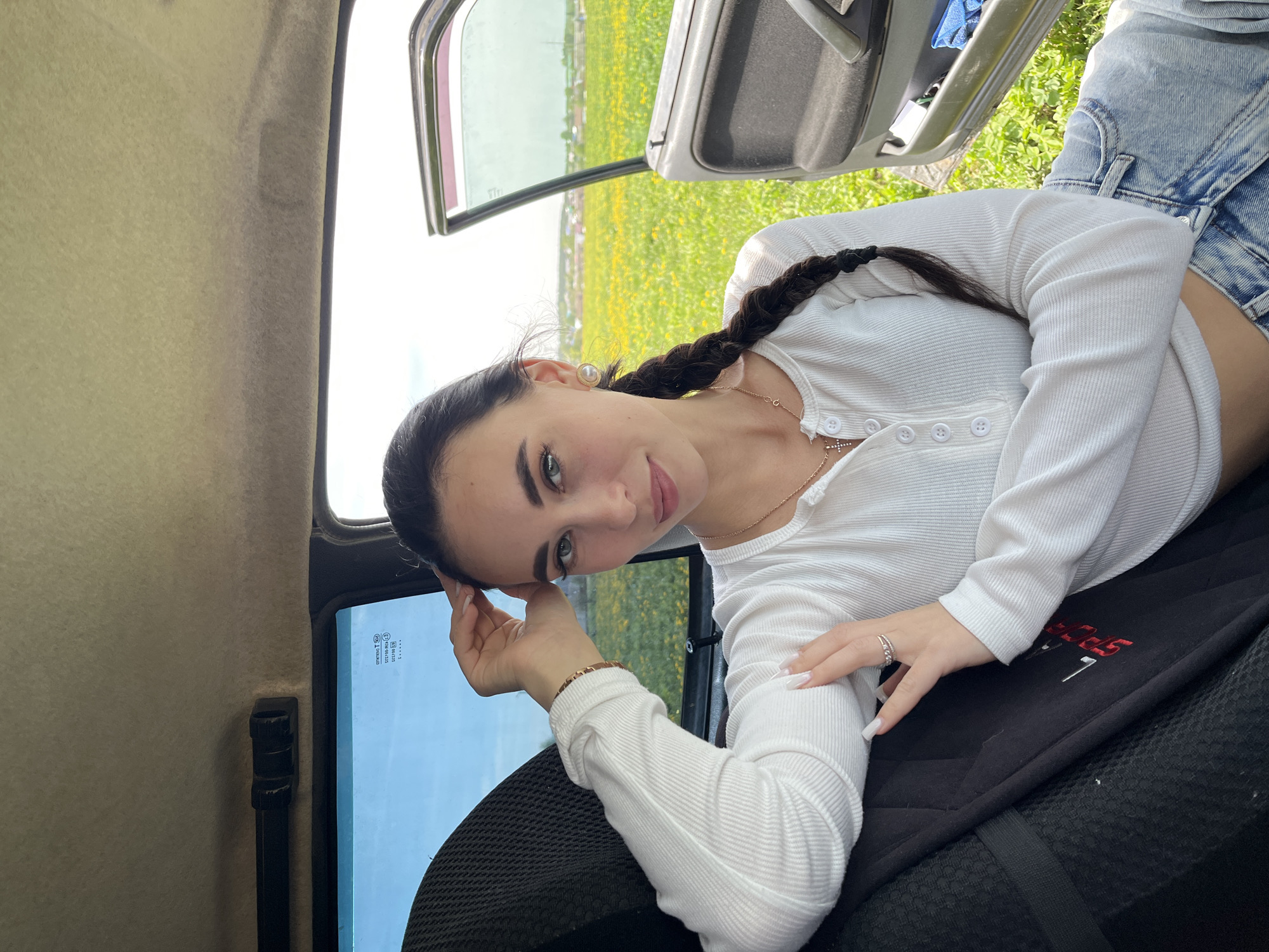 A young woman is reclining in the back of what appears to be a truck or similar vehicle, possibly in the vicinity of Фоки, Russia, given the presence of fields and vegetation visible through the open door. She is wearing a long-sleeved white top and light-wash denim shorts. Her hair is dark and styled in a braid. She is resting her head on her hand and looking towards the camera. The interior of the vehicle is visible, including the seat she is lying on and the door frame. The background shows a field with yellow flowers, suggesting it may be springtime.
