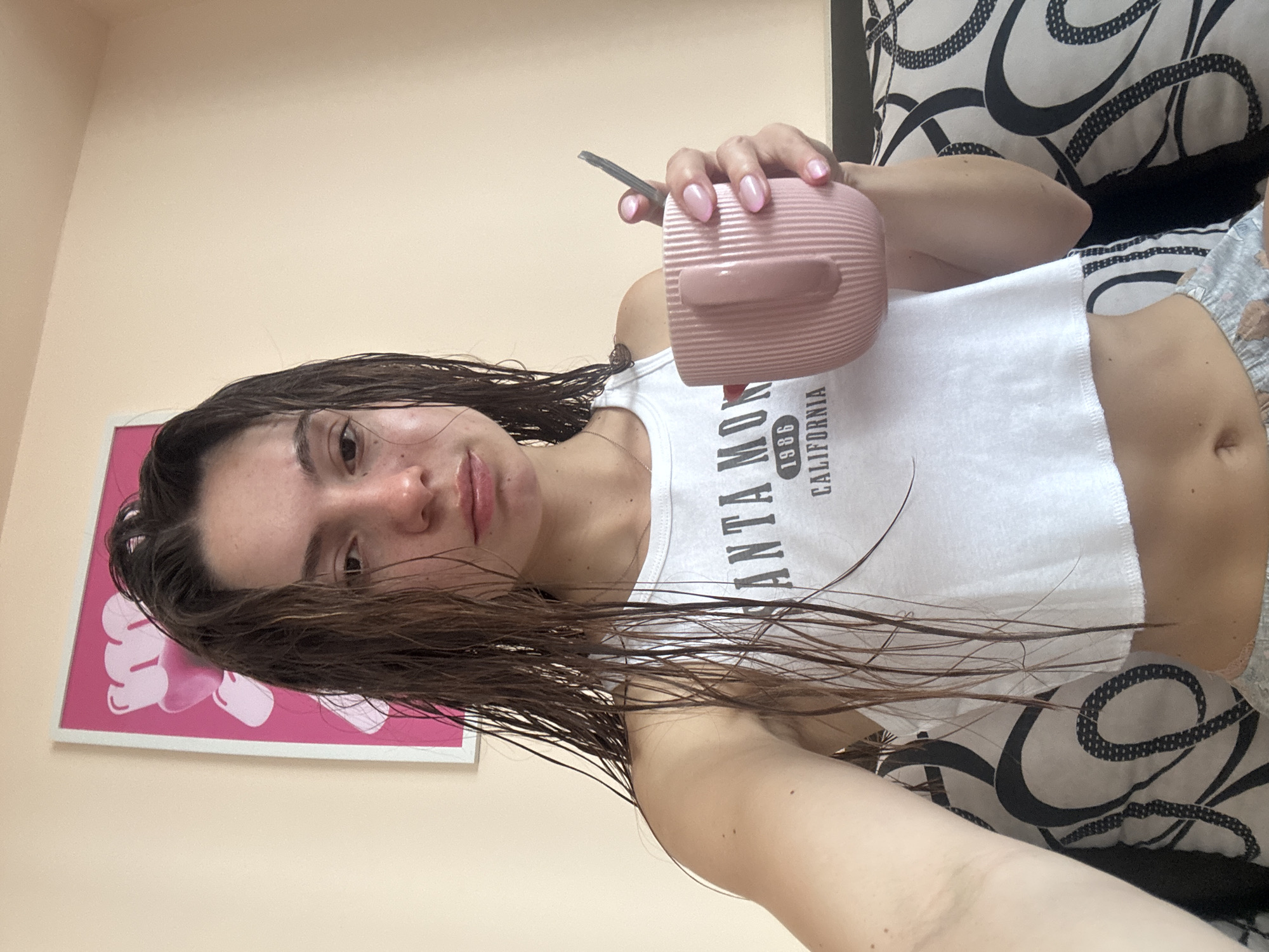 The image displays a selfie of a young woman indoors. She has long, dark brown hair that appears wet or damp, and a neutral, calm expression. She looks directly at the camera, showing a natural, possibly makeup-free face with visible freckles.
She is wearing a white sleeveless crop top with the text "SANTA MONICA 1986 CALIFORNIA" printed on it, revealing her midriff. Her lower attire, partially visible, consists of patterned shorts or pajamas. Her left hand holds a pink, textured mug or thermos, which contains a metallic straw or stirrer. Her fingernails are painted with light pink polish.
The background shows a light, pale peach or beige colored wall. On the left, a framed picture with abstract pink and white shapes (resembling hearts or bubbles) is visible. To the right, a black and white patterned pillow or bedding with swirling designs can be seen. The setting suggests a casual, personal space, likely a bedroom, in Varna, Bulgaria.