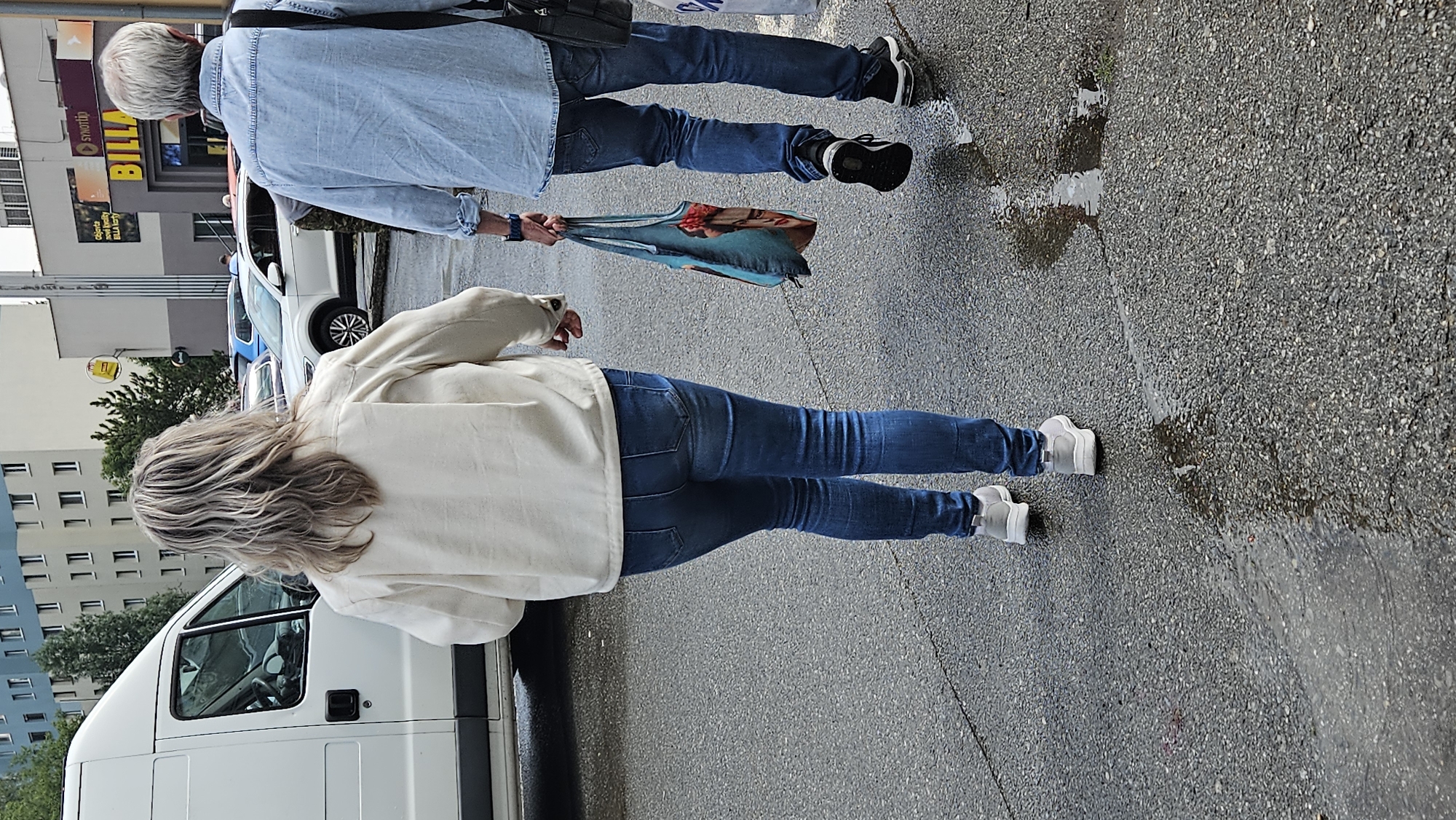 Two individuals are walking on a wet asphalt surface in the city of Prešov, Slovakia. The person in front, viewed from behind, has blonde hair and is wearing a cream-colored jacket and blue jeans. They are wearing white sneakers. The person behind them has gray hair and is wearing a blue denim shirt and blue jeans. They are carrying a patterned tote bag in their right hand and a black bag over their left shoulder. In the background, a white van is parked on the left, and a building with a yellow "BILLA" sign is visible on the right. The environment appears to be an outdoor urban setting, possibly a parking lot or street, with some trees and other vehicles present.