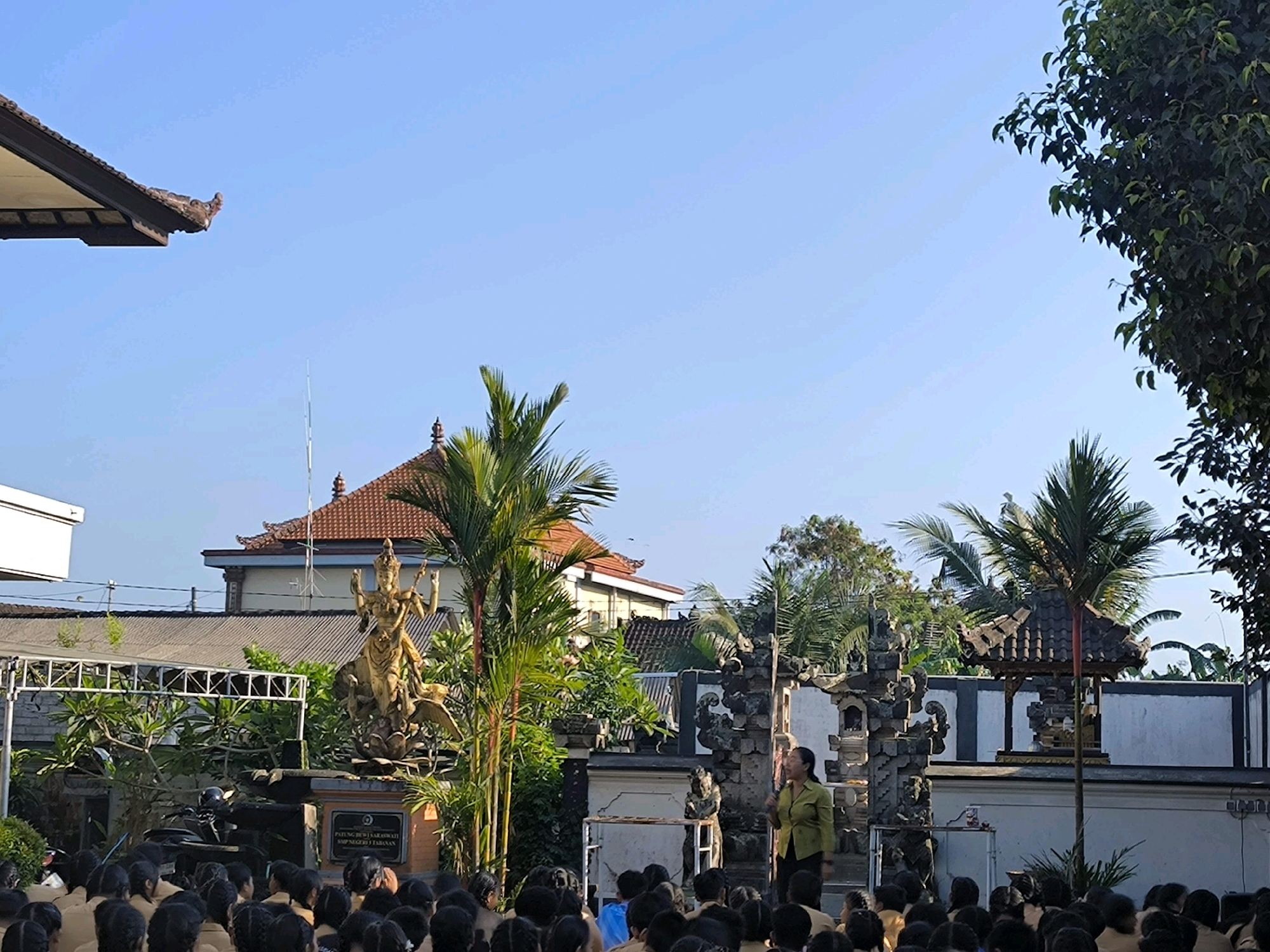 The image depicts an outdoor scene, likely in Tabanan, Indonesia, featuring a group of students attentively watching a woman in a lime green top and dark pants who appears to be speaking. In the background, a traditional Balinese temple gate structure is visible, adorned with intricate stone carvings. To the left of the gate, a golden statue of a multi-armed deity is prominently displayed, standing on a lotus flower. Palm trees and lush greenery surround the area, suggesting a tropical climate. The sky is a clear, bright blue, indicating a sunny day. The students are seated, facing the speaker, some wearing uniforms. The overall impression is of an educational or cultural event taking place in a visually rich, traditional setting.