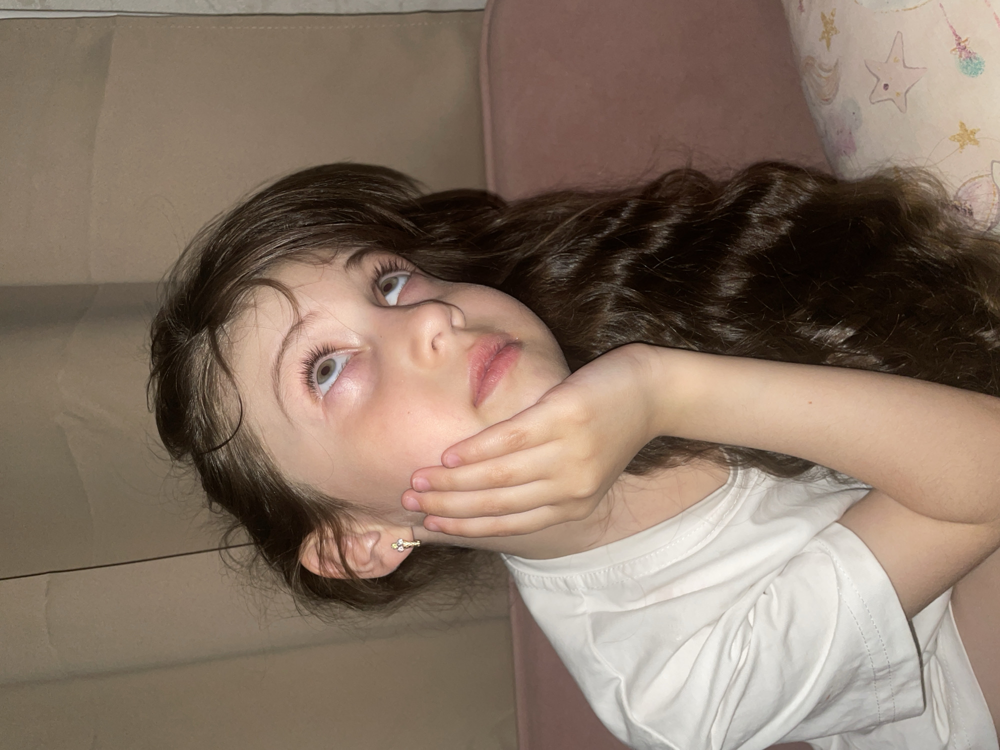 A young girl with long, wavy dark brown hair is depicted lying down, her head supported by her hand, as she gazes upwards. Her light-colored eyes are wide open, with long eyelashes, conveying a thoughtful or curious expression. She is wearing a simple white short-sleeved t-shirt. On her visible ear, she has a small, golden earring with what appears to be tiny decorative stones.
The setting appears to be indoors, likely in a relaxed environment such as a living room or bedroom. The background behind her head consists of a plain light brown or beige upholstered surface, possibly a sofa or a headboard. To her right, a soft pink surface is visible, and further right, a white pillow with light-colored patterns, including stars and other whimsical designs, adds a touch of childhood decor. The lighting suggests an indoor environment, possibly with artificial light, but a specific time of day is not clearly discernible. There is no visible text in the image.
While the location context is given as Izhevsk, Russia, no specific visual cues in the image itself directly indicate this geographical location. The scene focuses intimately on the girl's face and upper body, capturing a moment of quiet contemplation.