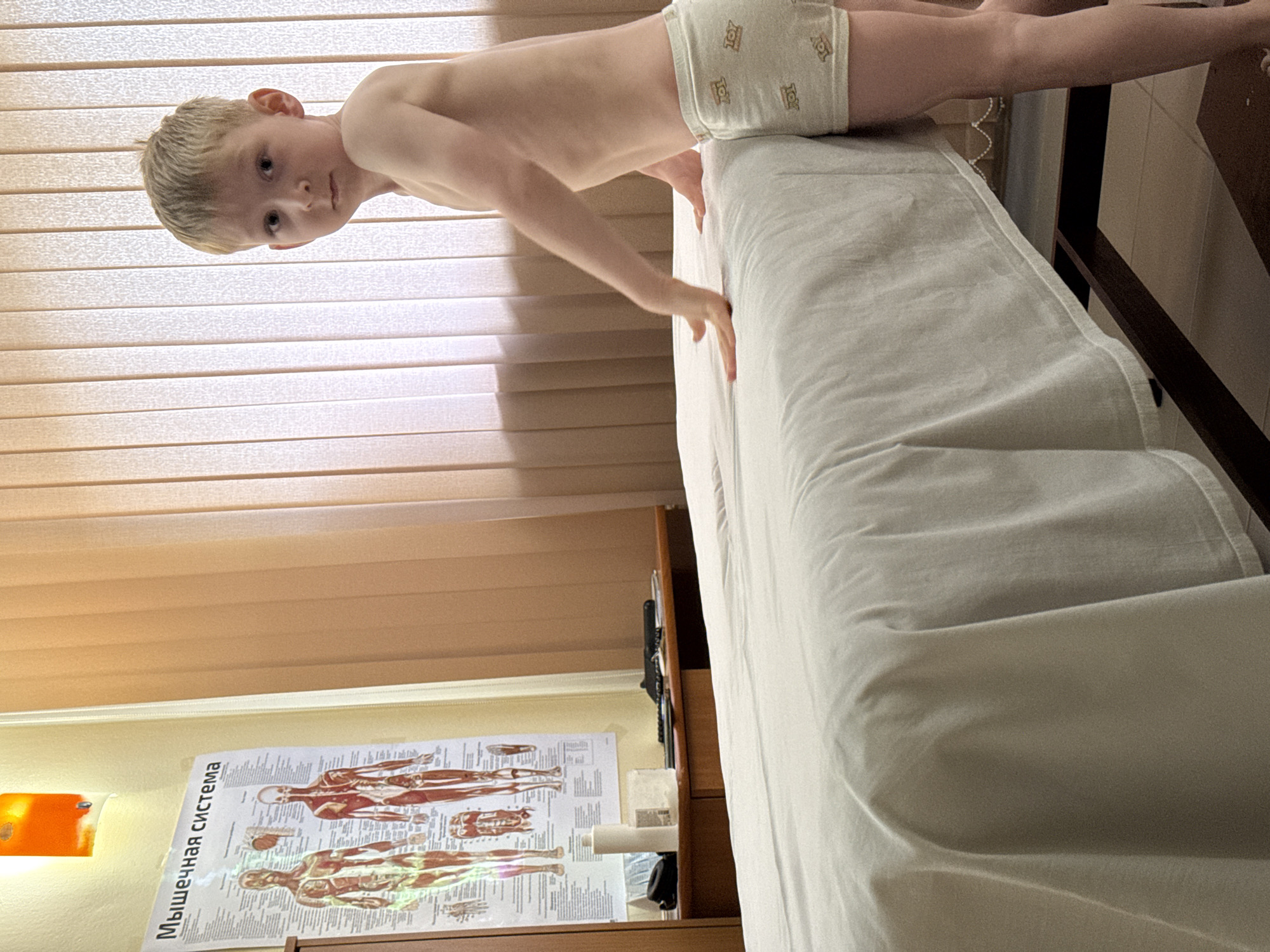 A blonde child wearing underwear with "Toy Story" characters is partially visible on a massage table covered with a white sheet. The room features light-brown window blinds and a poster displaying the human muscular system in Russian ("Мышечная система"). A wooden cabinet is positioned near the massage table. The location is likely a massage room or clinic in Chaikovsky, Russia, based on the poster language.