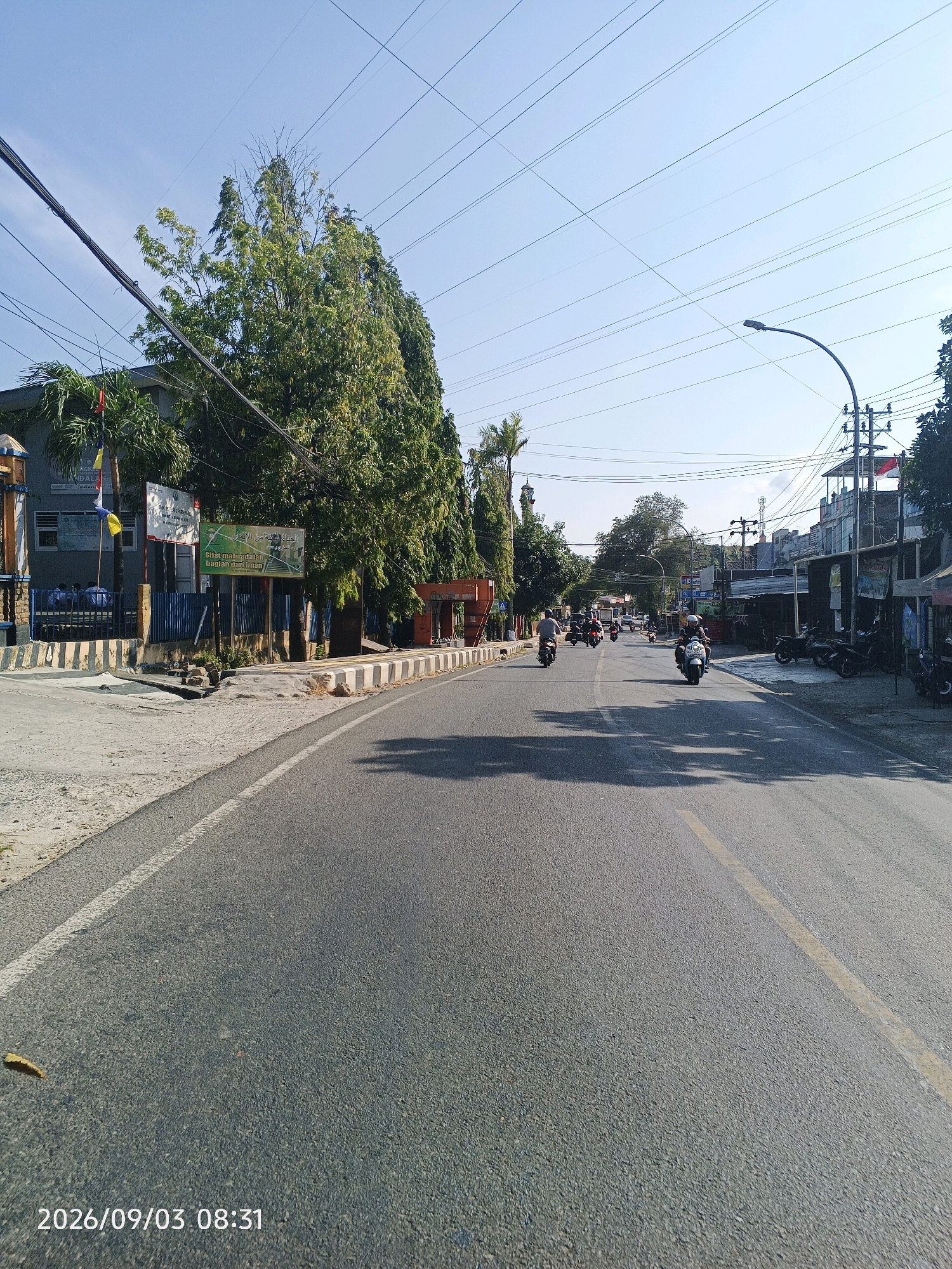 The image captures a street scene in Pare-Pare, Indonesia, on September 3, 2026, at 08:31. The viewpoint is from a moving vehicle, looking down a paved road lined with buildings and trees. Several motorcycles are visible, with riders navigating the street, indicating active local transportation. The buildings on the left appear to be part of an educational institution or government facility, with signage in Indonesian and Arabic scripts. The right side of the road features shops and businesses. Overhead, numerous power lines crisscross the clear blue sky, a common feature in Indonesian urban landscapes. The overall impression is of a typical morning commute in a bustling Indonesian city.
