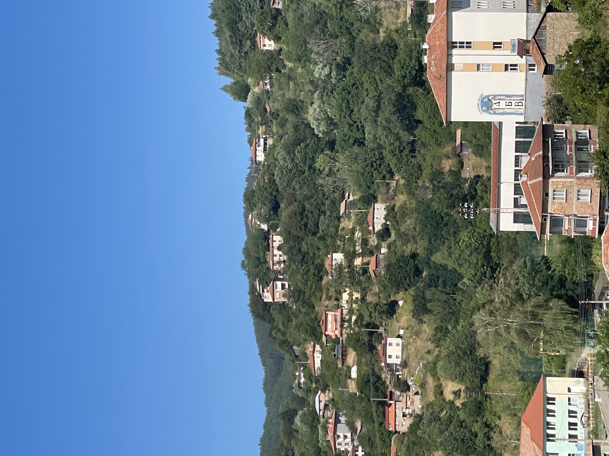 The image depicts a scenic view of the village of Davidkovo, Bulgaria, nestled on a hillside under a clear blue sky. The village is characterized by numerous houses with red-tiled roofs scattered across the lush green slopes, interspersed with dense trees. In the foreground, a modern building with a white facade and orange accents stands out. This building features a mural on its side depicting two figures and Cyrillic lettering. To the left of this building, another structure with a light blue and beige facade and numerous windows is partially visible. The overall scene is tranquil and pastoral, showcasing a traditional Balkan village environment.
