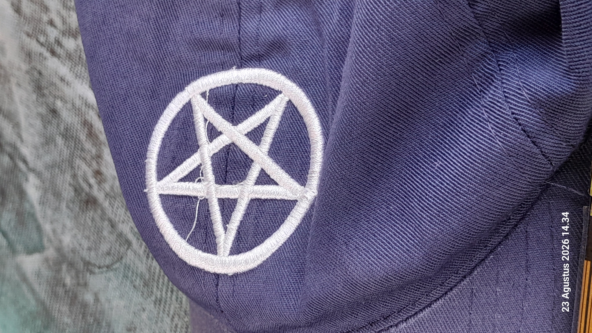 A close-up, medium-angle, outdoor shot shows a blue fabric with a white embroidered pentagram. The fabric is likely part of a hat or cap, as indicated by the curved seam visible in the lower left. The pentagram is centered on the fabric, with each point of the star reaching towards the outer edge of a circle. The background is out of focus, but appears to be a textured, possibly fabric surface in muted greens and grays. The lighting is even, with no harsh shadows. The image is timestamped "23 Agustus 2026 14.34" in the lower left corner. No people or specific landmarks of Gemaharjo, Indonesia are visible.
