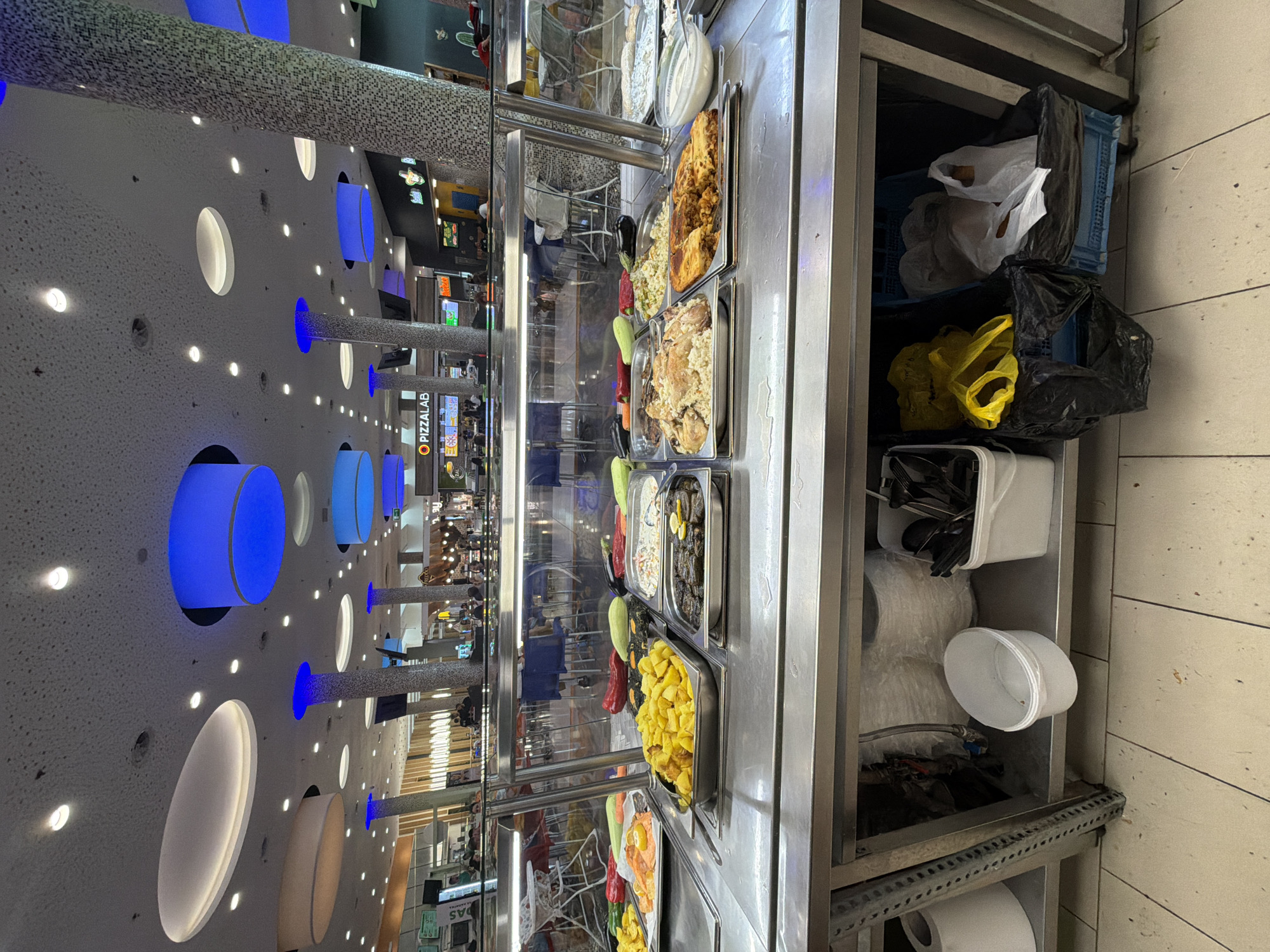 This image, captured indoors in Plovdiv, Bulgaria, depicts a section of a modern food court or cafeteria. The perspective is rotated 90 degrees to the left, showing a food display counter on the right and extending into the dining area.
The main subject on the right is a stainless steel food display unit, presenting a variety of prepared dishes in metal trays. Visible items include what appear to be baked potatoes or fried yellow vegetables, roasted chicken pieces alongside rice or grains, a green salad or mixed vegetables, a creamy white sauce or dip, and another tray with fish or dark roasted items. Decorative elements like whole zucchini and bell peppers are placed between some trays. Beneath the counter, a storage area holds blue plastic crates, assorted plastic bags (white, yellow, black, some containing bread), white containers holding cutlery, and rolls of paper towels.
The ceiling is a notable feature, white with numerous circular cutouts. Some circles are illuminated with vibrant blue lights, while others emit a warm white glow. Small, bright white spotlights are scattered across the ceiling. Several pillars, covered in a mosaic pattern of small white and blue tiles, ascend through the space, some also featuring blue lighting.
In the background, a broader view of the food court reveals tables, chairs, and other food stalls. Blurry figures of people can be seen sitting or moving around, suggesting an active environment. A clear sign for "O PIZZALAB" is visible among the background establishments. The setting appears brightly lit and functional, typical of a busy indoor dining area, possibly within a shopping mall.