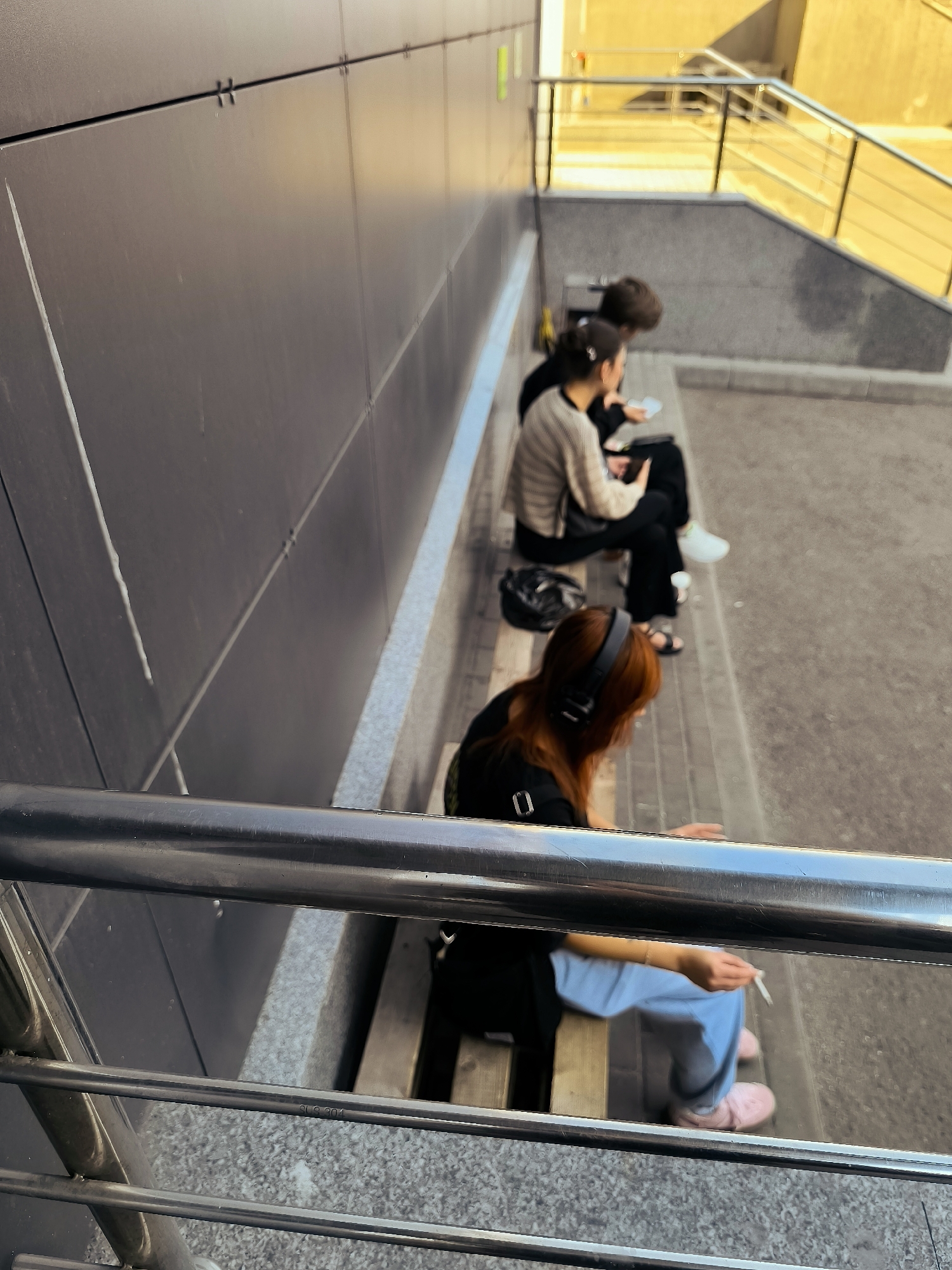 The image depicts an outdoor scene, possibly a public seating area near a stairway. In the foreground, a metal railing runs horizontally across the frame. Behind the railing, several individuals are seated on a bench.
The closest individual, with reddish-brown hair, is wearing a black top and light blue pants. They have headphones on and are holding a cigarette in their right hand. Their left hand is resting on their lap. To their left, on the bench, is a black bag.
Further back, another individual, possibly a woman with dark hair tied back, is wearing a beige and white striped sweater and black pants. They are holding a phone or similar device in their hands. Beside them, another person with dark hair is partially visible, also appearing to be looking at a device.
The background shows a grey, textured wall and a stairway with yellow handrails, suggesting an urban or public infrastructure setting. The lighting suggests it is daytime. The specific location in the city of Qarağandı city administration, Kazakhstan, is not discernible from the visual elements in the image itself.