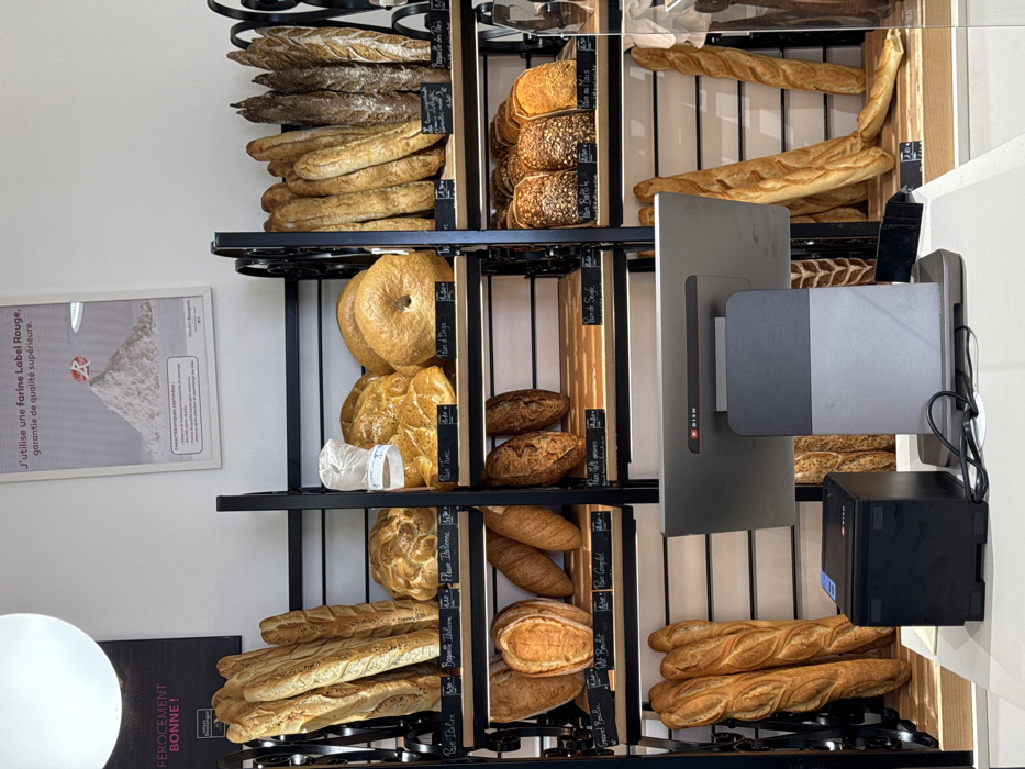 This image captures the interior of a boulangerie (bakery) in Asnières-sur-Seine, France, showcasing a wide selection of bread products. The scene is dominated by multiple black metal shelving units with light wooden shelves, displaying an array of freshly baked bread.
On these shelves, various types of bread are arranged: numerous long baguettes of varying crust colors (light to dark), round loaves, oval loaves, braided bread, and distinctive bagels. Many items are labeled with small black price tags in French, indicating types such as "Pain Italien," "Baguette Italienne," "Pain Turc," "Pain d'Orge," "Grand Boule," "Petit Boulot," "Pain Complet," "Pain Battu graines," "Pain de Seigle," "Pain Battu," "Pain de Maïs," and "Baguette des Prés."
In the foreground, a modern point-of-sale (POS) system is visible on a white counter. It consists of a grey monitor with a "DISH" logo, angled away from the viewer, and a black POS printer located to its left. Electrical cords are visible near the base of the monitor.
The walls are white. On the upper right wall, a framed poster features a large pile of white flour with a red "R" emblem and French text stating, "J'utilise une farine Label Rouge, garantie de qualité supérieure." A partially visible sign on the upper left wall reads "FÉROCEMENT BONNE!" A large, spherical white light fixture hangs from the ceiling in the upper left. No individuals are present in the image. The environment appears clean and well-lit.