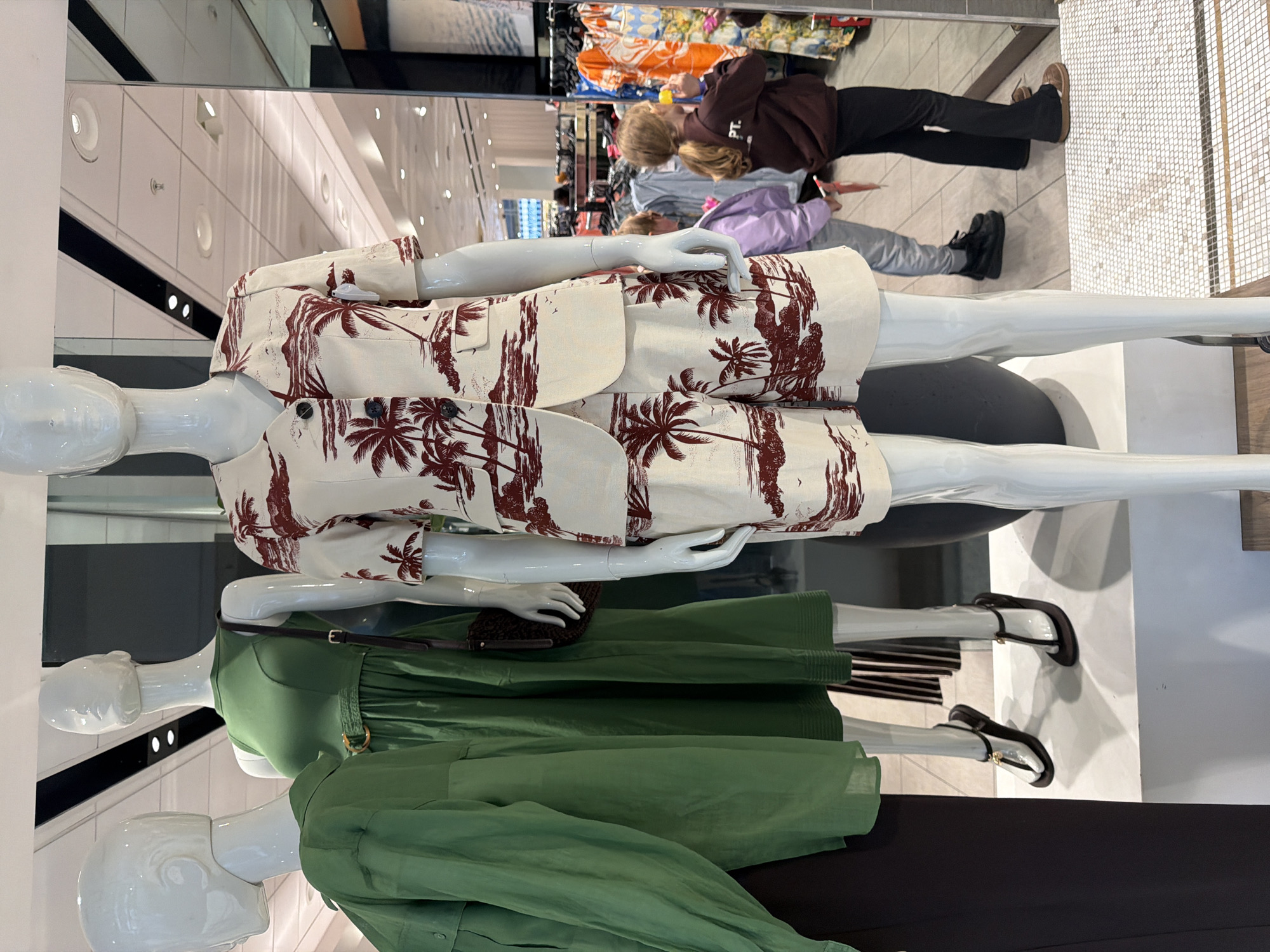 The image depicts a retail display inside a store, likely a clothing shop. Three white mannequins are arranged in a row. The first mannequin is dressed in a printed beige co-ord set featuring a tropical tree motif in reddish-brown. The second mannequin wears a midi-length green dress with a brown belt. The third mannequin is dressed in a long green shirt and black bottoms.
In the background, one can discern the reflection of shoppers in a mirror. The clothing racks in the background are stocked with apparel. The architecture of the store includes white walls and recessed lighting. Based on the style and brands visible, the store could be located in London, United Kingdom.